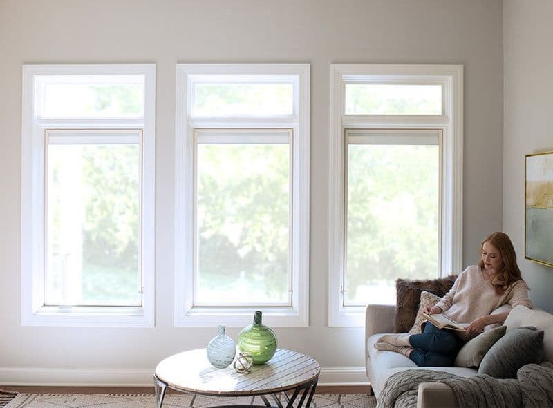 Woman reading a book on a sofa in a brightly lit room featuring three large white picture windows with transom lights above them.