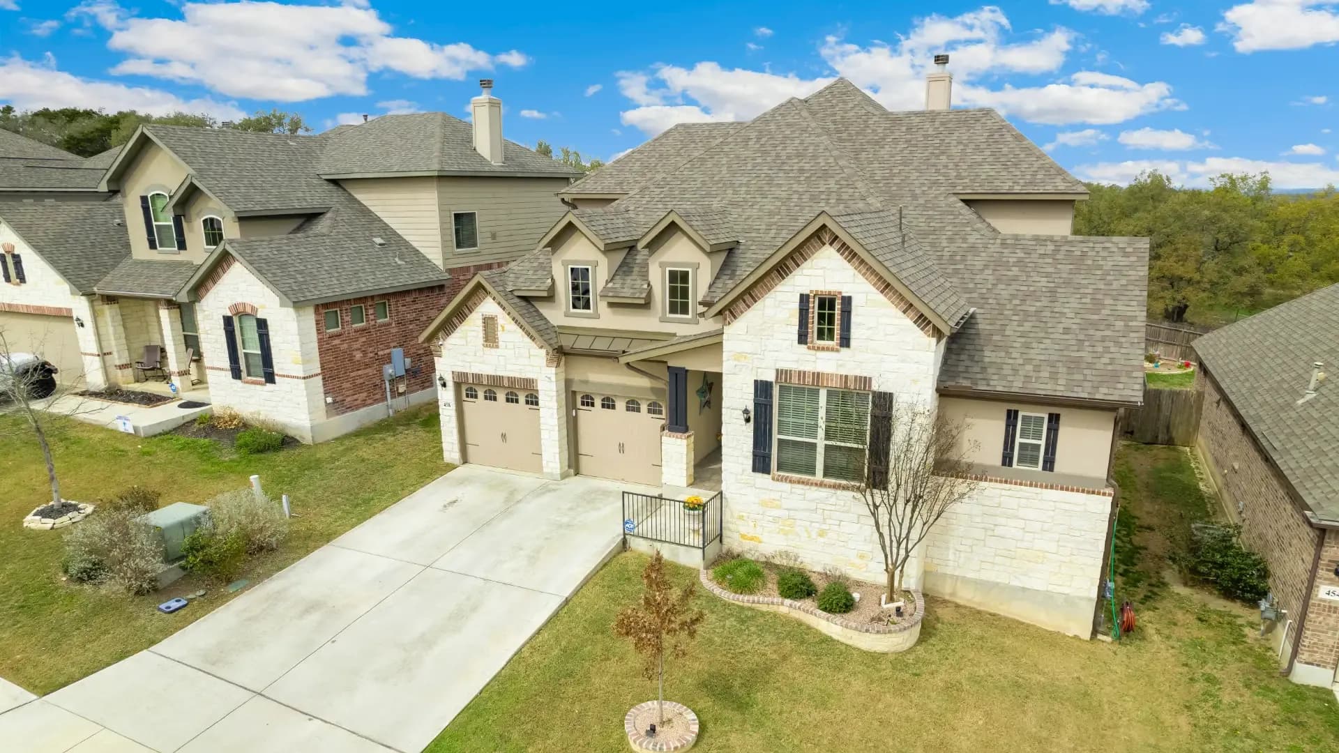 Aerial view of a suburban house with a stone facade and a three-car garage. The home features a manicured lawn, and the neighborhood is lined with similar homes under a bright blue sky.