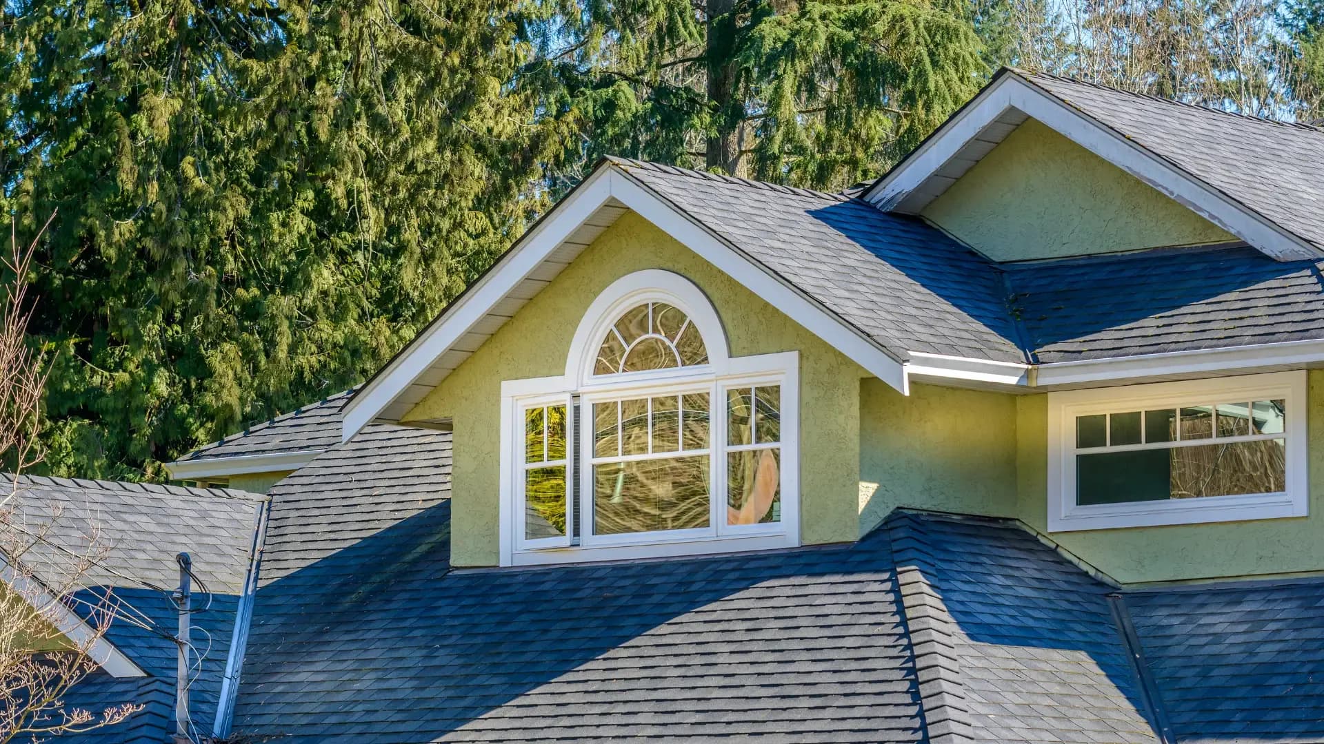 Arched window with decorative grilles set into the green stucco gabled upper story of a house with dark roof shingles.