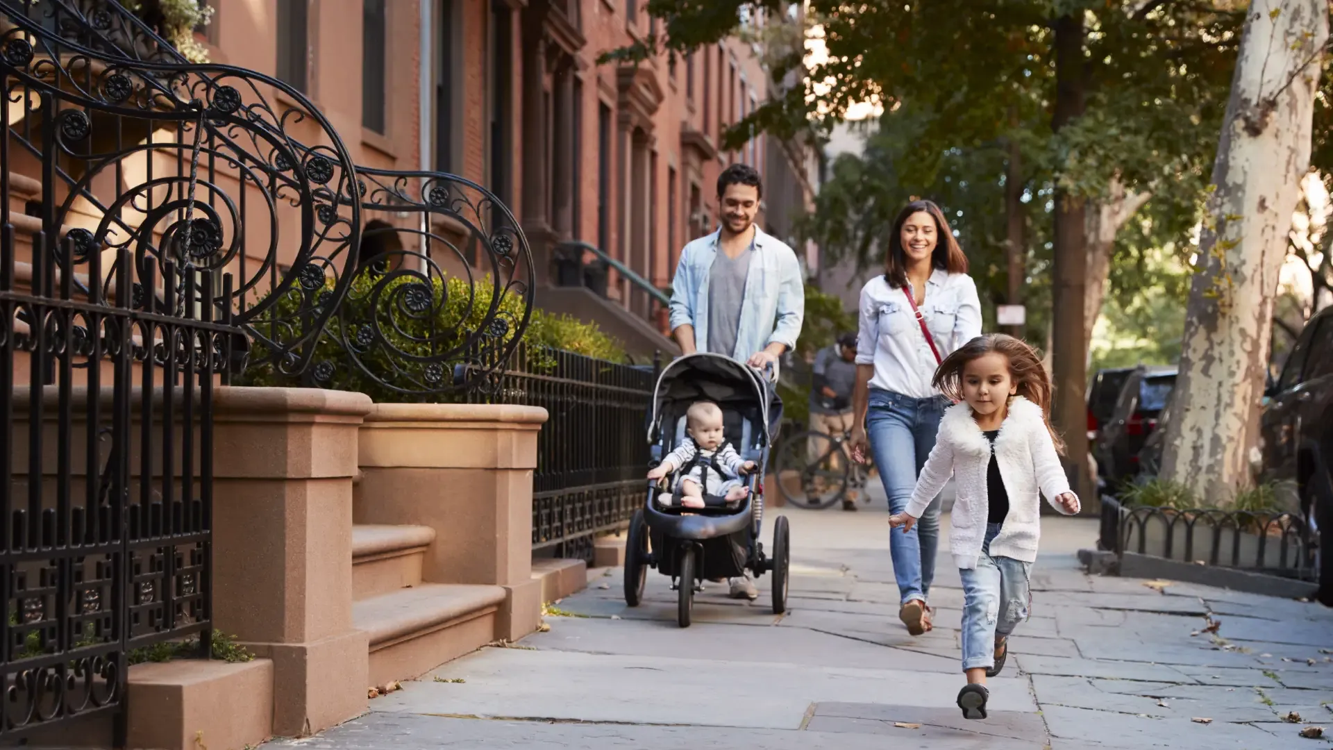 Family with a toddler in a stroller taking a walk down a residential sidewalk lined with brownstone buildings.