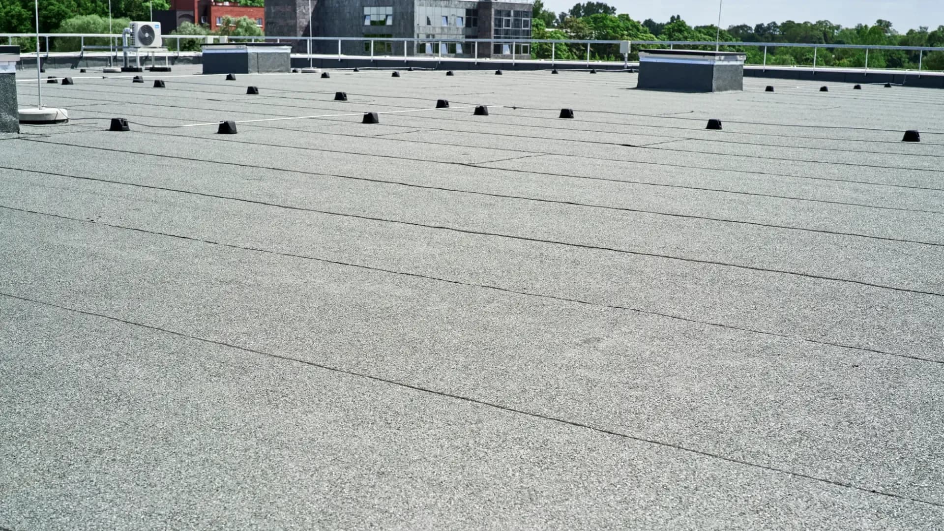 Wide perspective view of a large commercial flat rooftop covered with a light grey rolled asphalt protective membrane and black support blocks under a clear sky.
