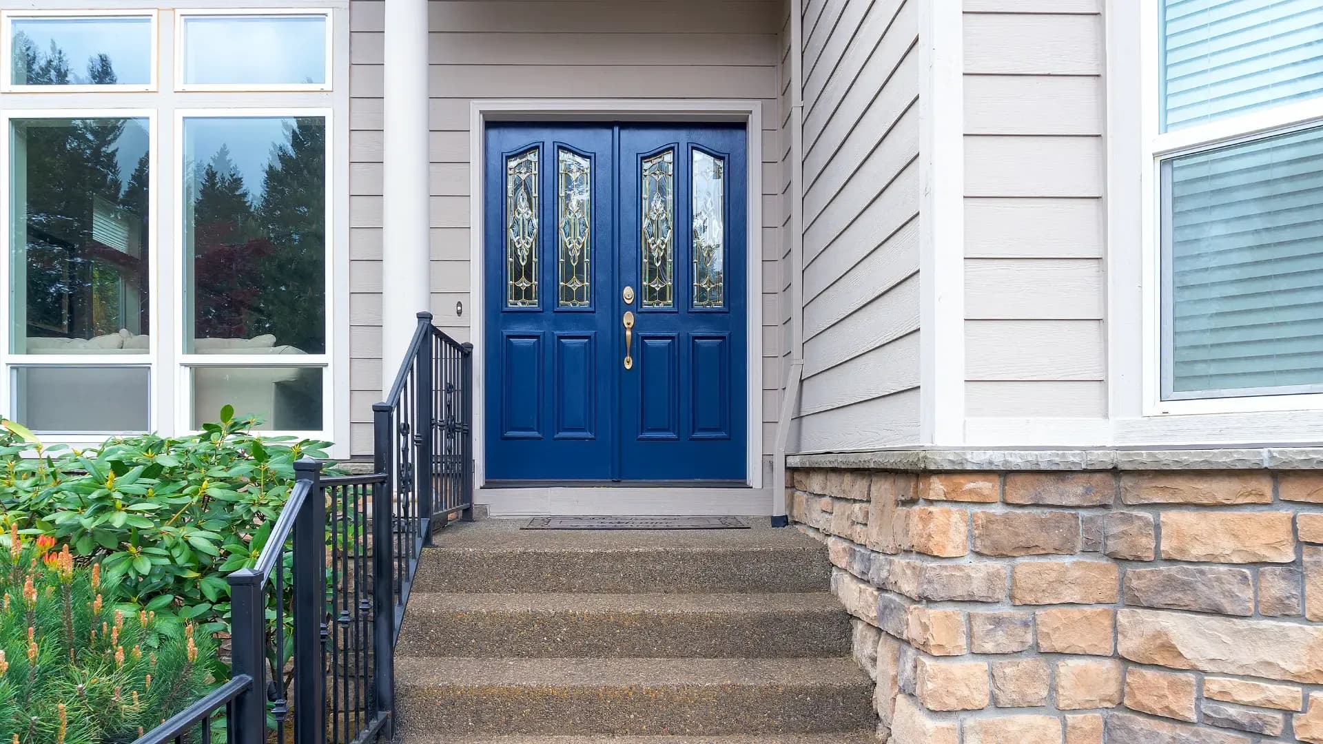Navy blue double front entry doors with ornate decorative glass inserts, accessible via concrete steps with a black wrought iron railing.