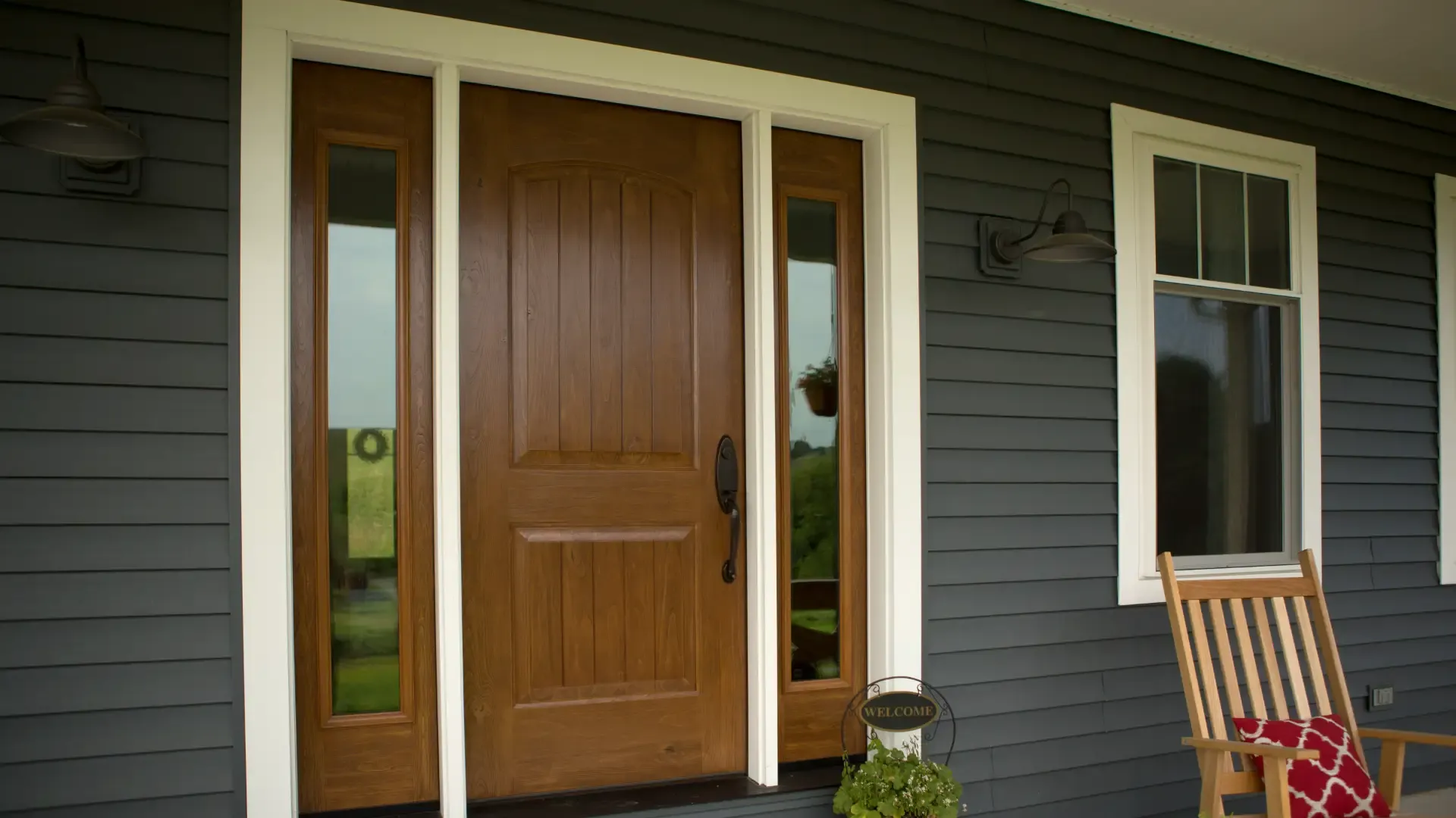 Exterior view of a dark grey horizontal lap siding porch featuring a medium-toned wood front entry door with full-length sidelights, bright white trim casing, a white-framed single-hung window, and a wooden rocking chair with a red patterned pillow.