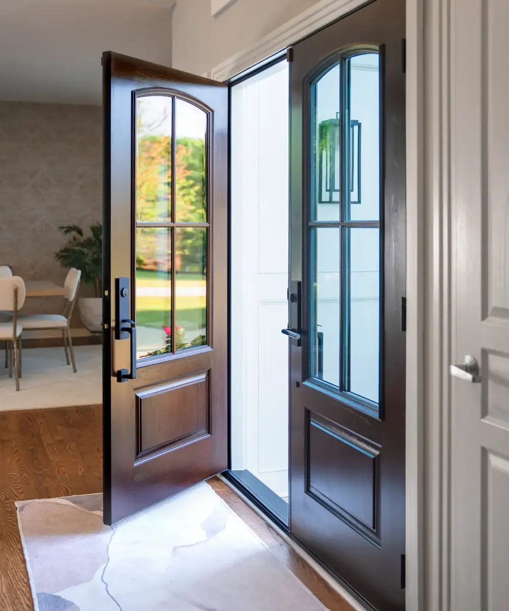 Interior view of open dark hazelnut French entry doors featuring arched four-lite glass inserts and modern black hardware, looking out onto a bright entryway and showing medium-toned hardwood floors inside.