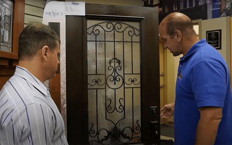 Two men looking at a dark wood entry door featuring decorative wrought iron and frosted glass.