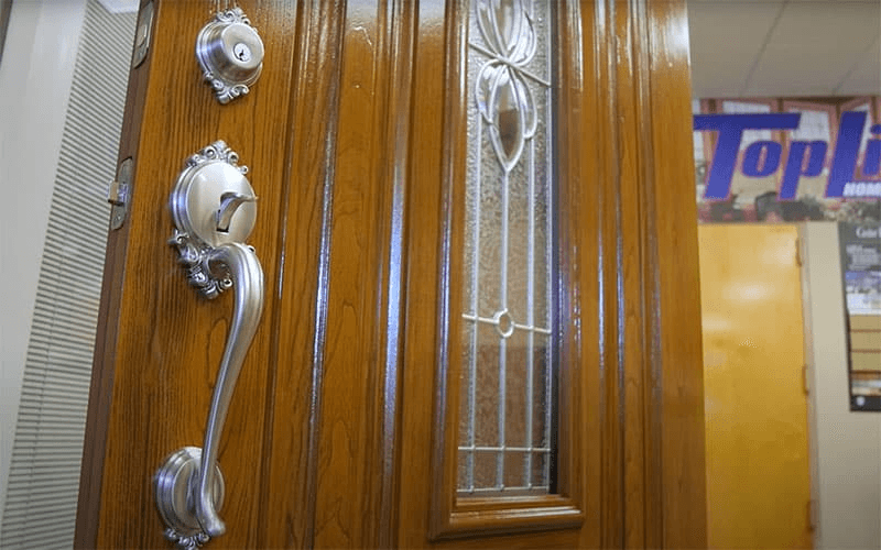 Wood-grain front door featuring an ornate brushed nickel handle, deadbolt, and a decorative frosted glass window insert.