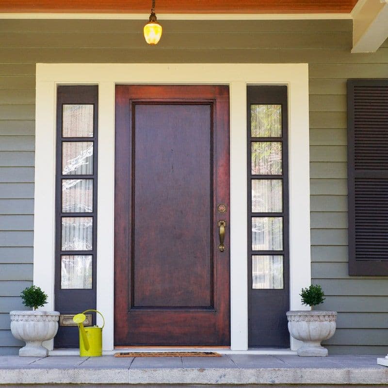 A brown wooden front door with glass panels on each side, flanked by potted plants and a yellow watering can. A warm porch light hangs above.