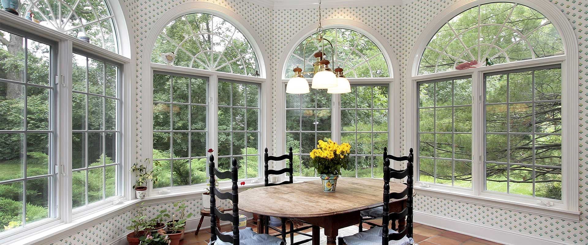 Sunroom dining area surrounded by large arched windows with grid patterns overlooking a lush green yard.