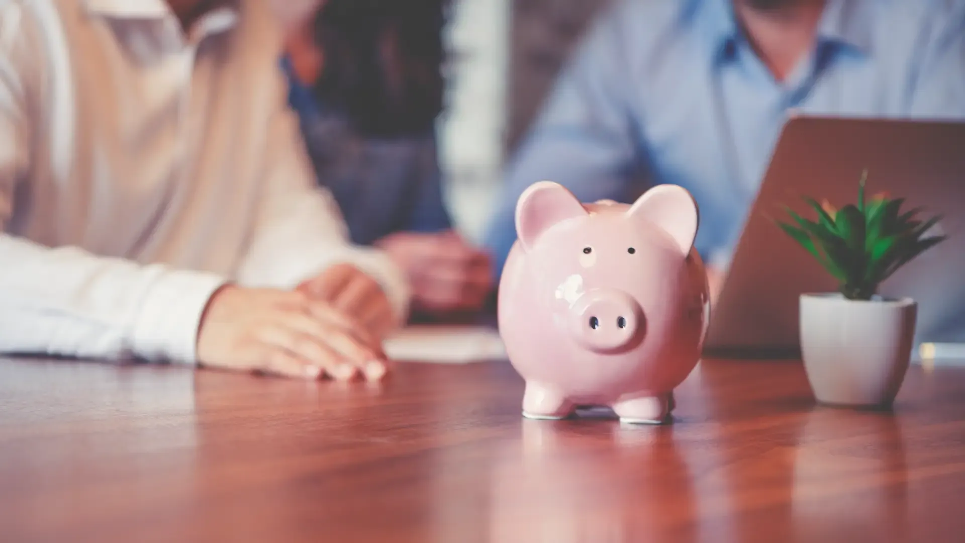Pink Piggy Bank on Wooden Desk (GettyImages-1127866568.webp) Close-up view of a glossy pink piggy bank standing prominently on a polished wooden desk, next to a small potted succulent and a laptop, with two people sitting blurred in the background.