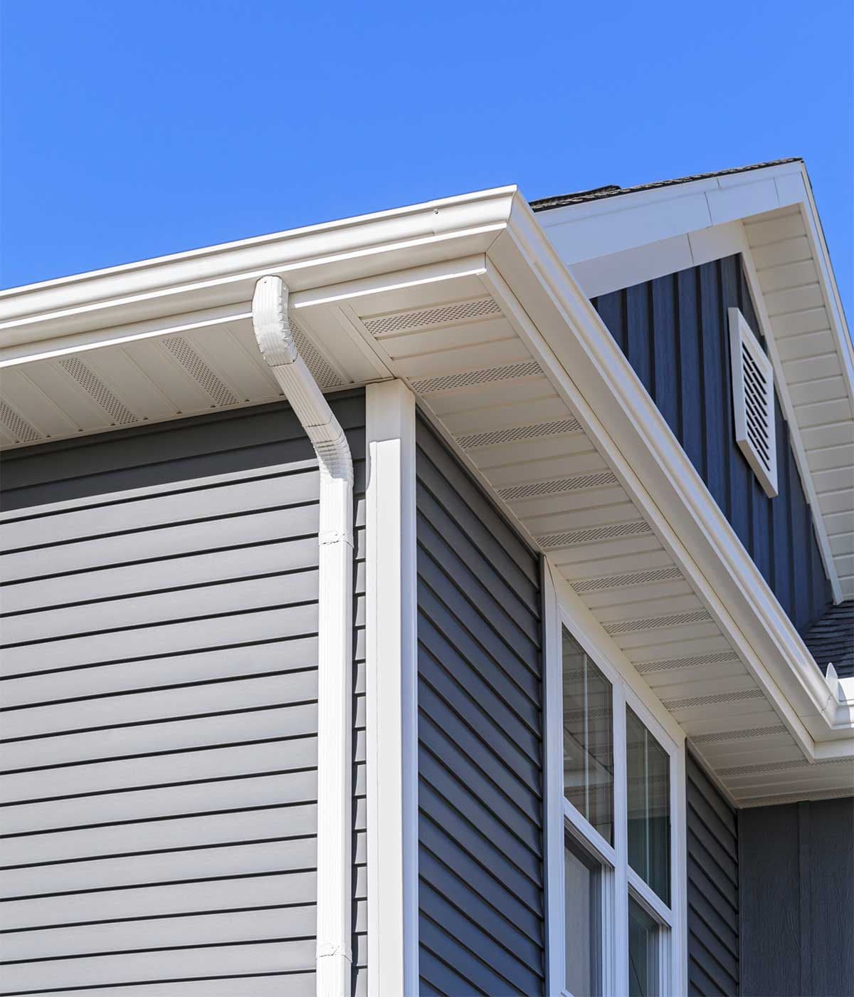 Low-angle exterior view of white seamless aluminum gutters, soffits, and a vertical downspout installed on a multi-story house with dark grey lap siding under a bright blue sky.