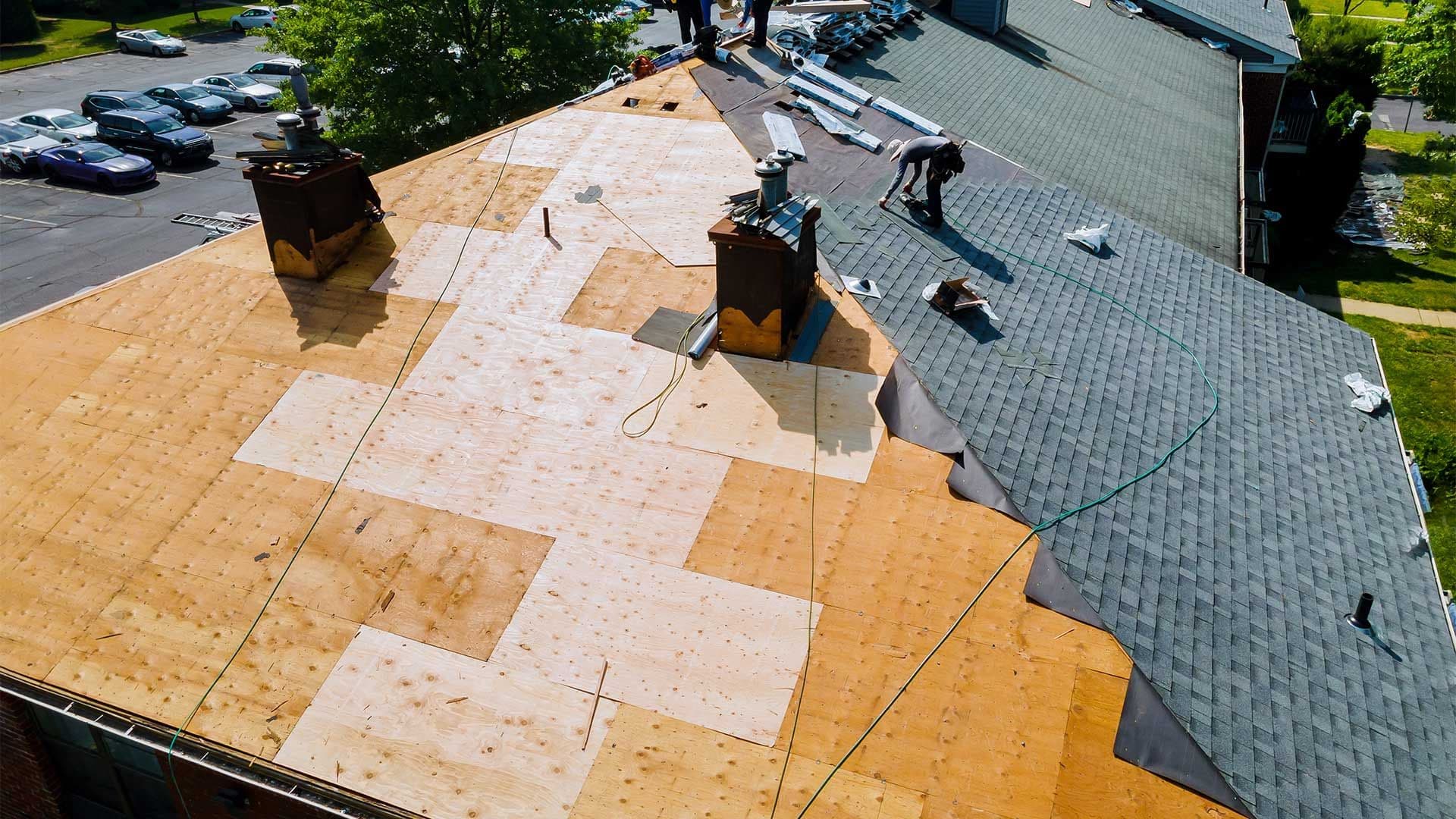 An aerial perspective showing a large roof undergoing replacement, featuring exposed plywood roof decking partially covered with black underlayment and newly laid grey asphalt shingles.