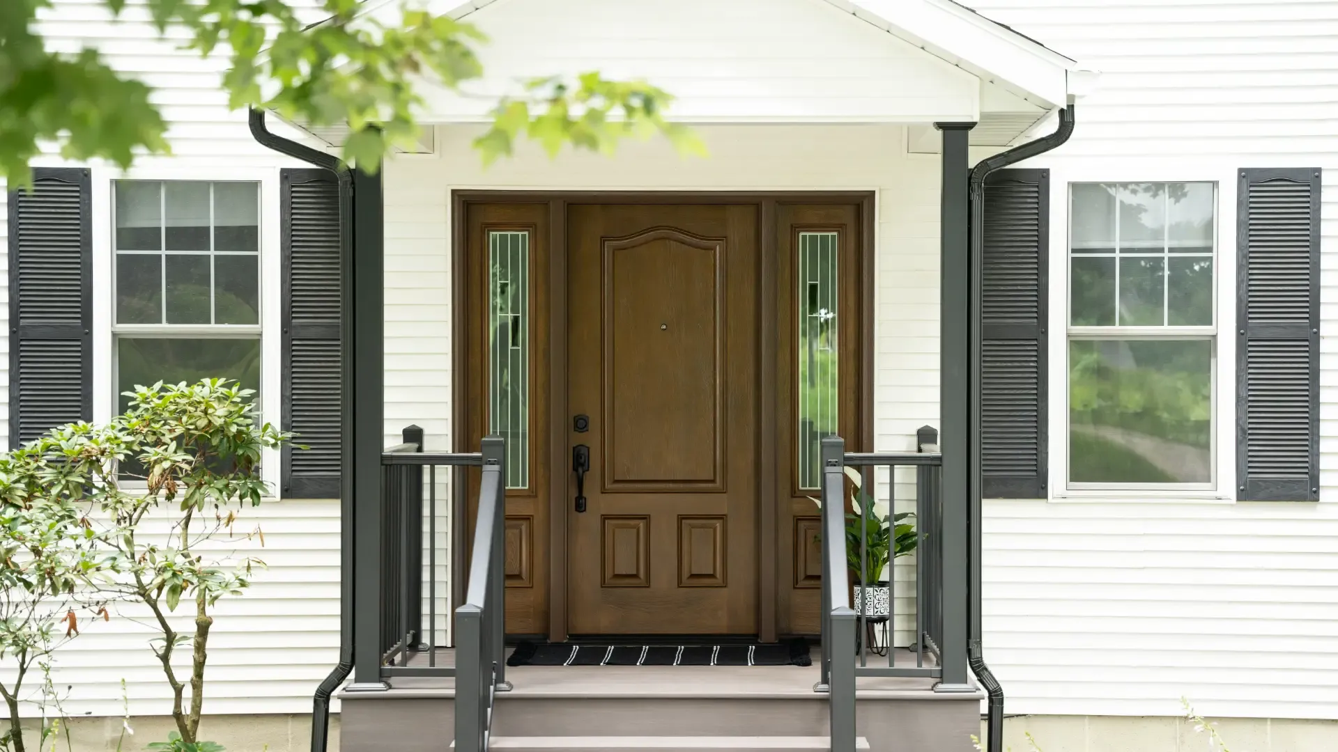 Exterior view of a home entryway featuring a dark brown wood front door with decorative paneling and two matching sidelights with narrow vertical glass inserts. The entry is framed by a white-clad portico porch supported by dark posts with matching dark railings and steps. On either side, white lap siding is accented by white-framed double-hung windows with colonial grilles and black shutters.