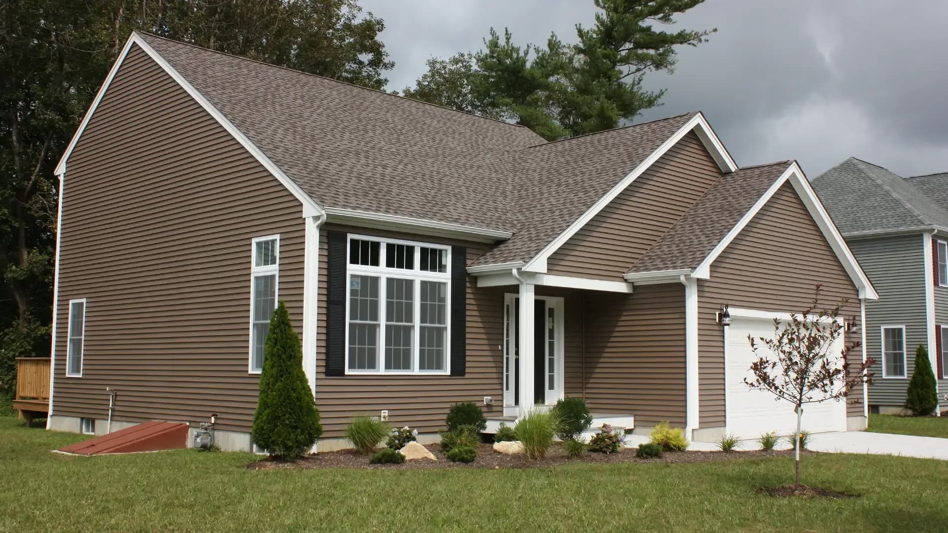 Exterior view of a modern single-story home clad in brown horizontal vinyl siding with white trim, featuring a multi-gable roofline, attached garage, white-framed grid windows with black shutters, and young front lawn landscaping.