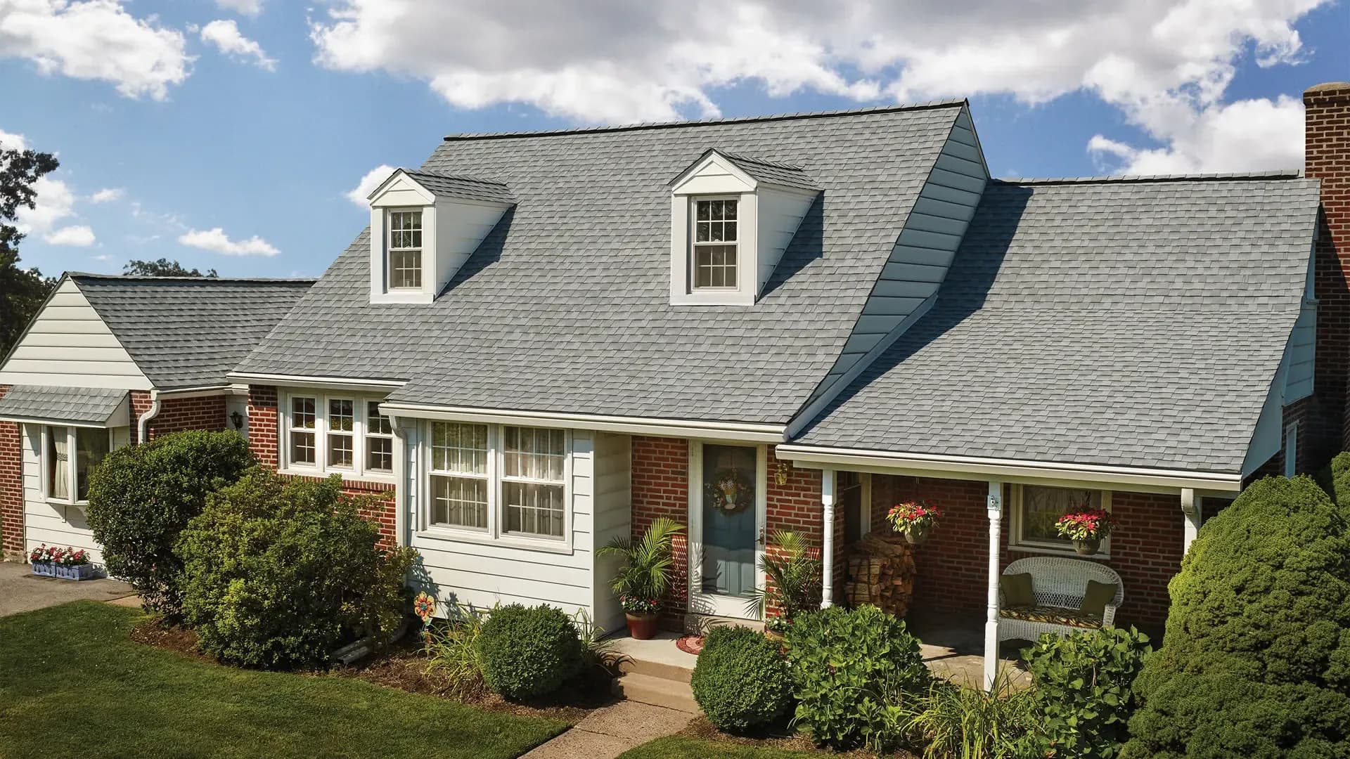 Exterior view of a multi-gabled brick Cape Cod home featuring GAF Timberline HD shingles in Fox Hollow Gray, white dormers, a covered front porch with hanging flower baskets, and lush green landscaping.