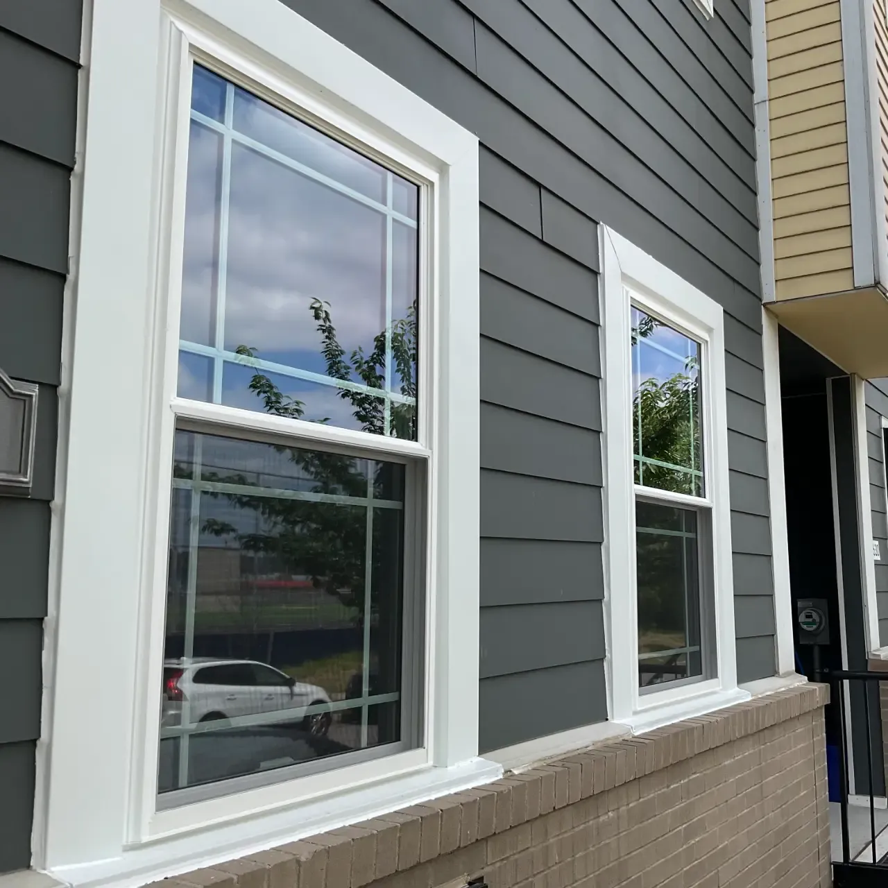 Two white double-hung windows with Prairie-style grids installed on a house with dark grey lap siding and a low brick accent wall.