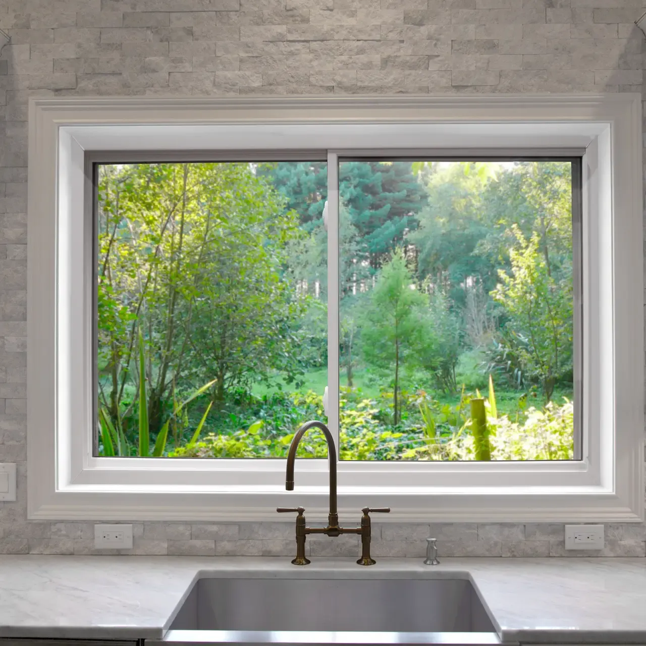 A white horizontal sliding window positioned directly above a kitchen sink, set against a light grey stacked stone backsplash. The window frames a lush, green view of trees and foliage outdoors, while a dark bronze gooseneck faucet is mounted on the white marble countertop in front of it.