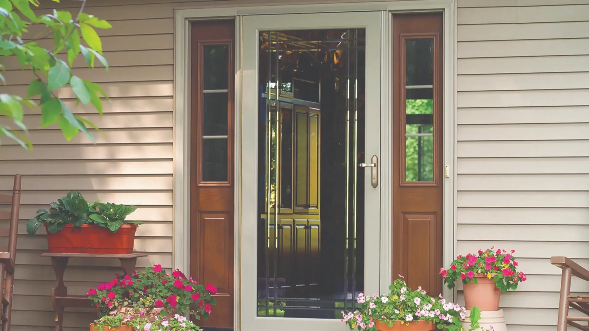 A glass front door with wooden panels, framed by two narrow windows. Potted flowers in vibrant pinks and reds add a welcoming touch to the entrance.