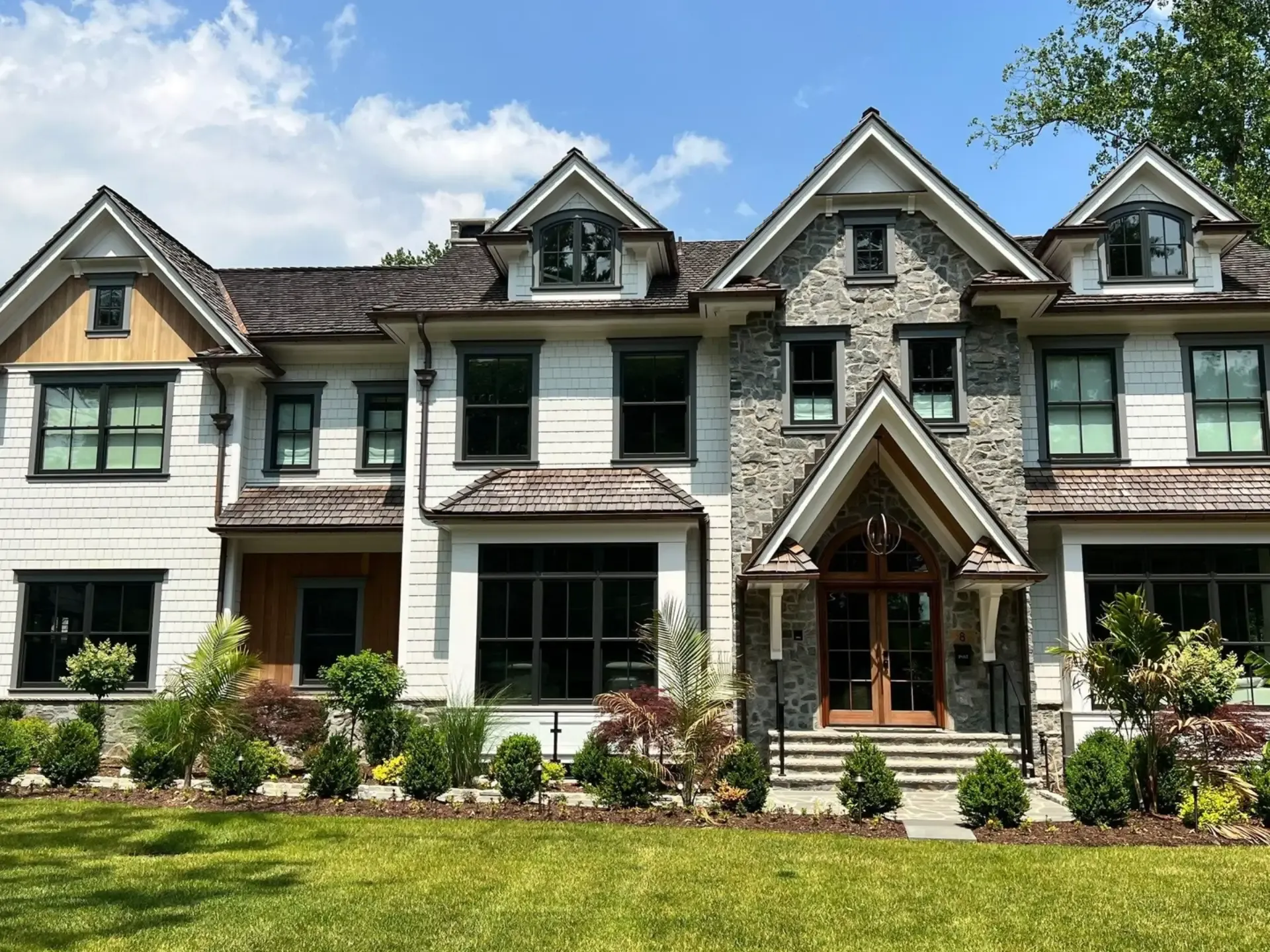 Exterior front view of a large two-story luxury craftsman-style home featuring white shake siding, gray stone masonry accents, dark gray framed windows, wood-paneled gables, and double arched wood front doors under a gabled stone entry porch with a green manicured lawn in the foreground.