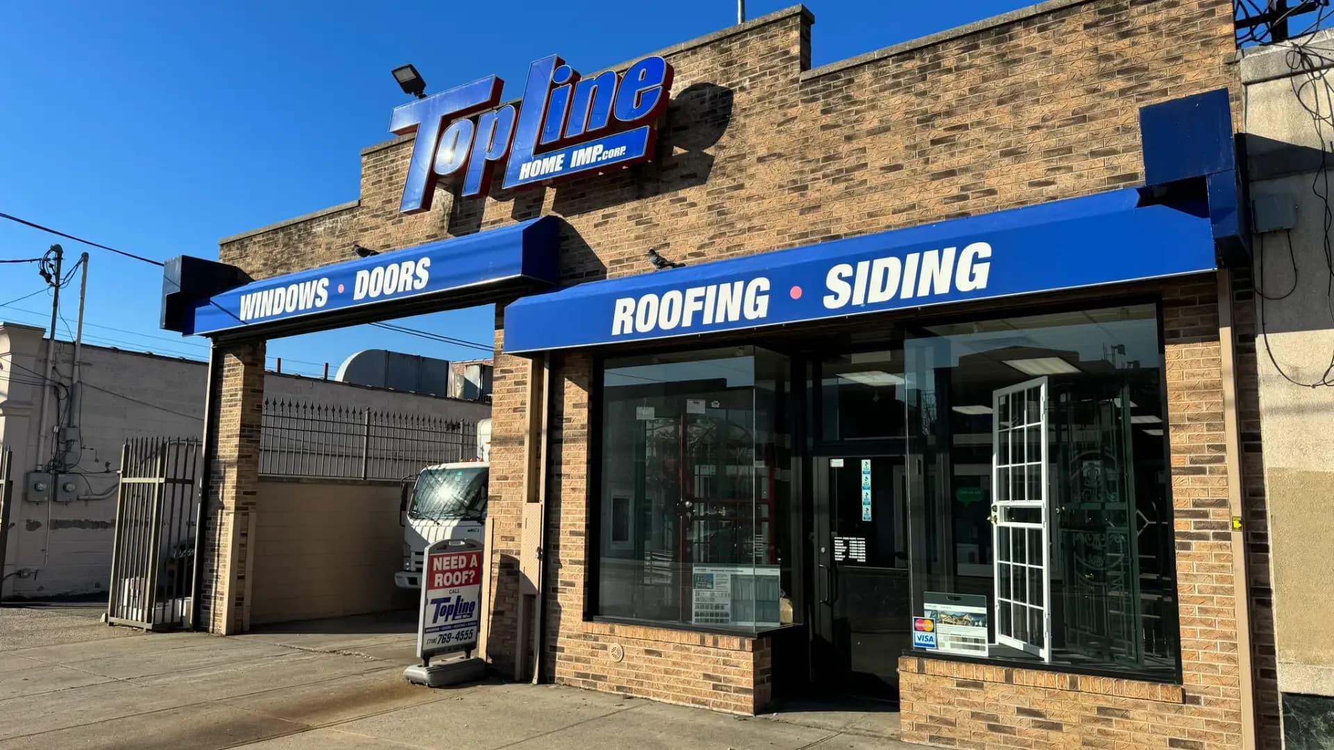 Bright daytime view of the Topline Home Improvement brick showroom featuring blue awnings that advertise windows, doors, roofing, and siding.