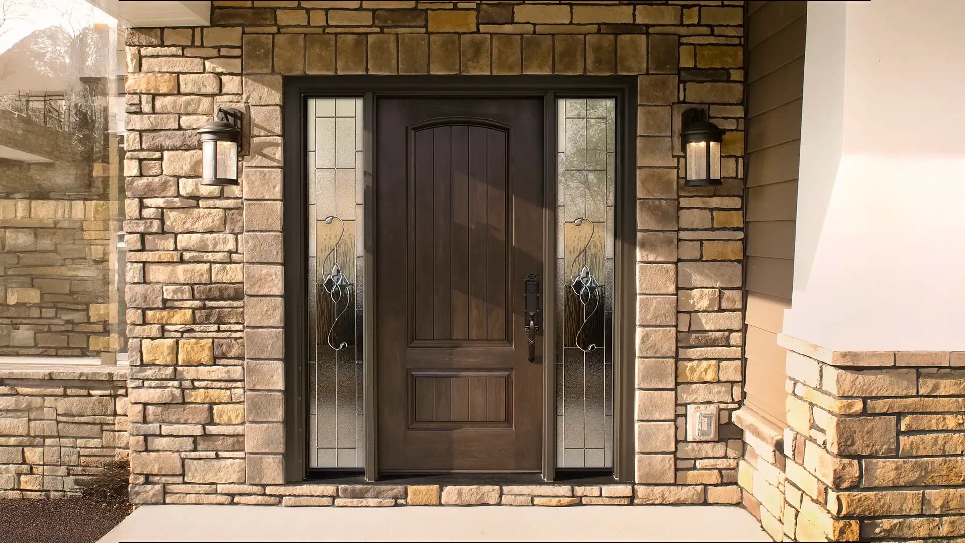 Elegant wooden front door with vertical glass panels on each side, framed by rustic stone walls. Two stylish lanterns add a welcoming touch.