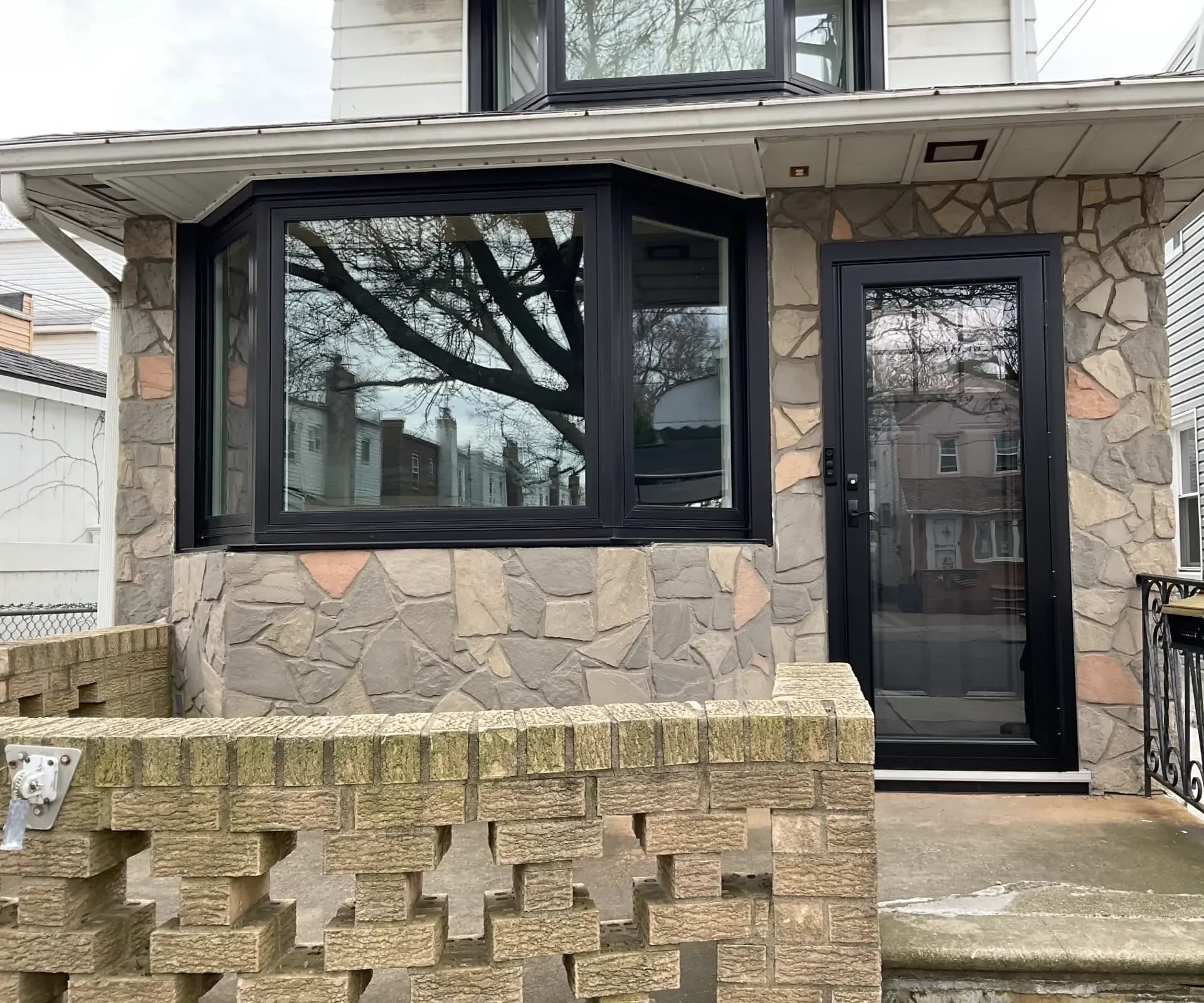 Black OKNA bay window and matching black glass storm door installed on a house facade featuring stone veneer siding and a brick porch wall.