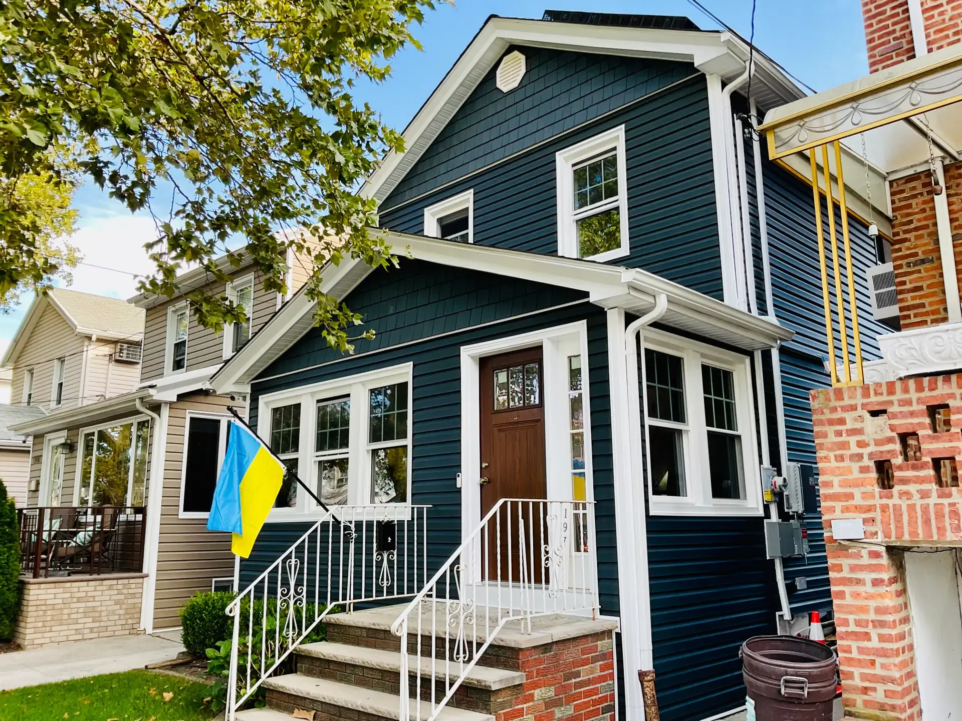 Exterior perspective view of a two-story house clad in dark navy blue siding with white trim, featuring a warm wood front door, white porch railings, and a blue-and-yellow Ukrainian flag mounted near the brick steps.