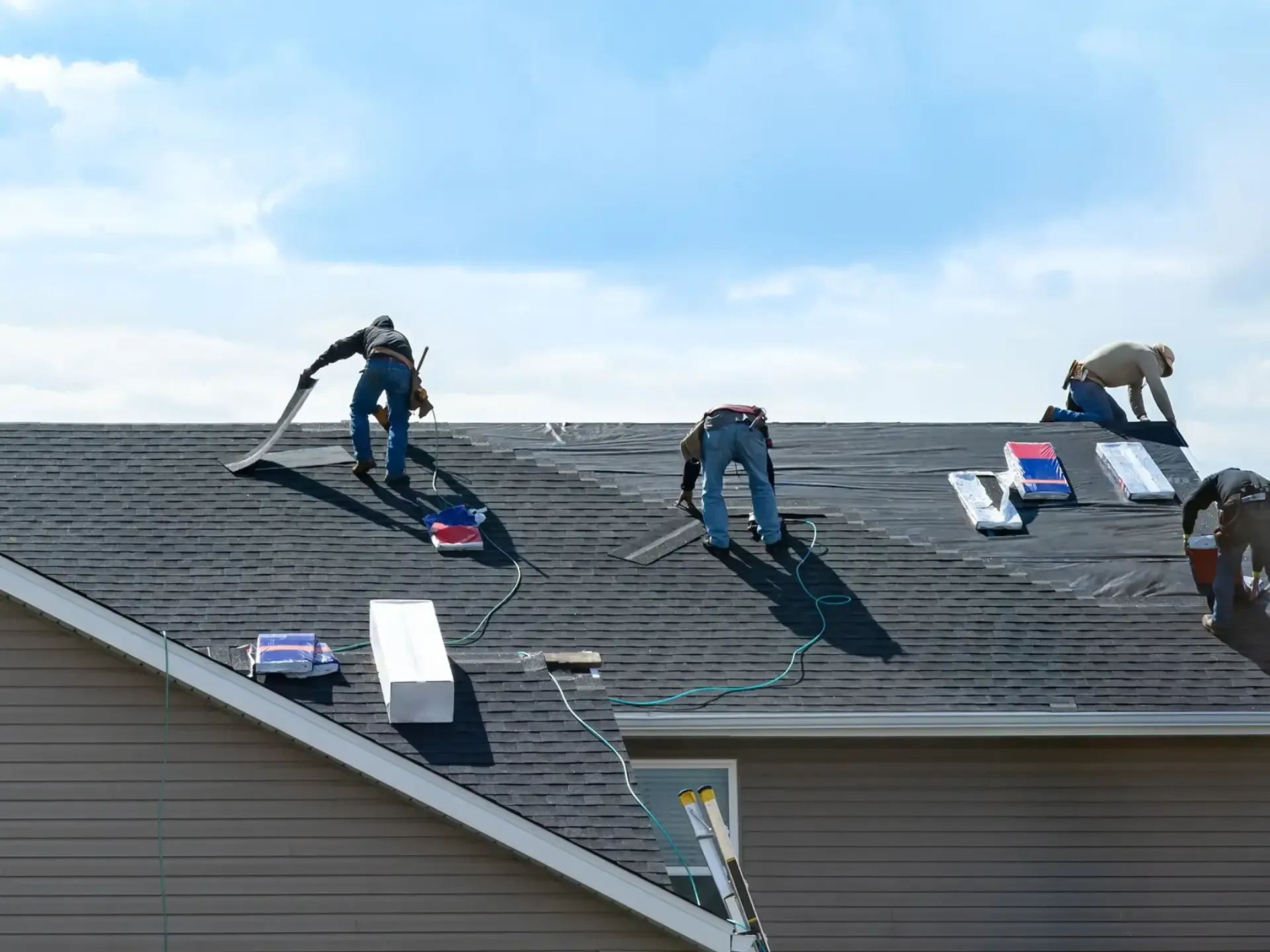 A wide exterior shot of a multi-person roofing crew actively working together to lay down black underlayment and install asphalt shingles across a large residential roof slope.