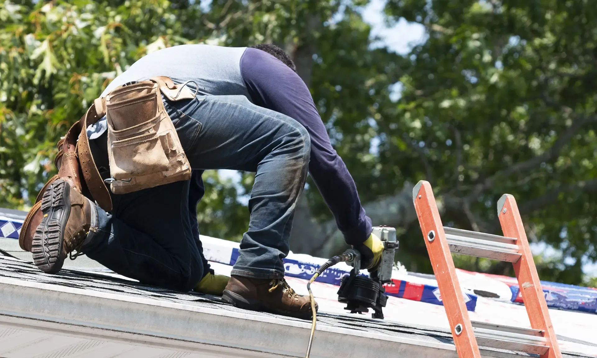 A close-up view of a professional contractor wearing a tool belt and work boots kneeling on a roof slope while securing asphalt shingles with a pneumatic coil nail gun.
