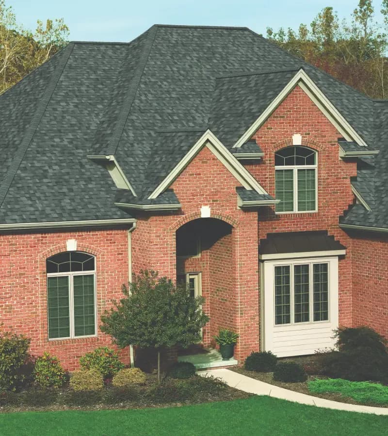 Estate Gray Architectural Shingle Roof on Red Brick Home (Owens Corning Duration® Premium Estate Gray Beauty Shot .webp) High-angle exterior view of a multi-gabled red brick home featuring Owens Corning Duration® Premium architectural shingles in Estate Gray, highlighted by arched top windows, a white bay window pop-out, and lush surrounding landscaping.