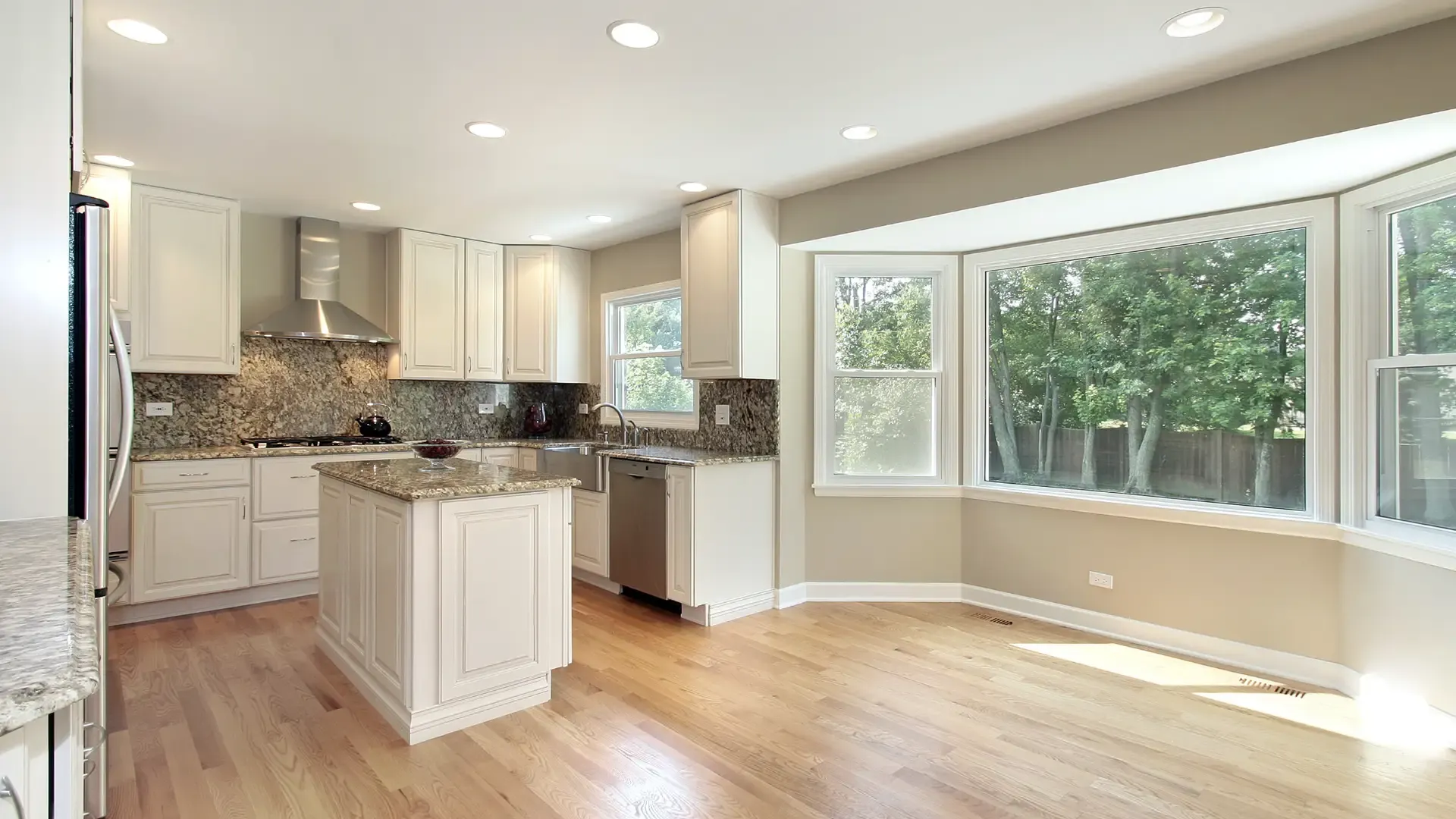 Bright kitchen with wooden floors, white cabinetry, and a granite countertop island. Stainless steel appliances and large windows with garden views.