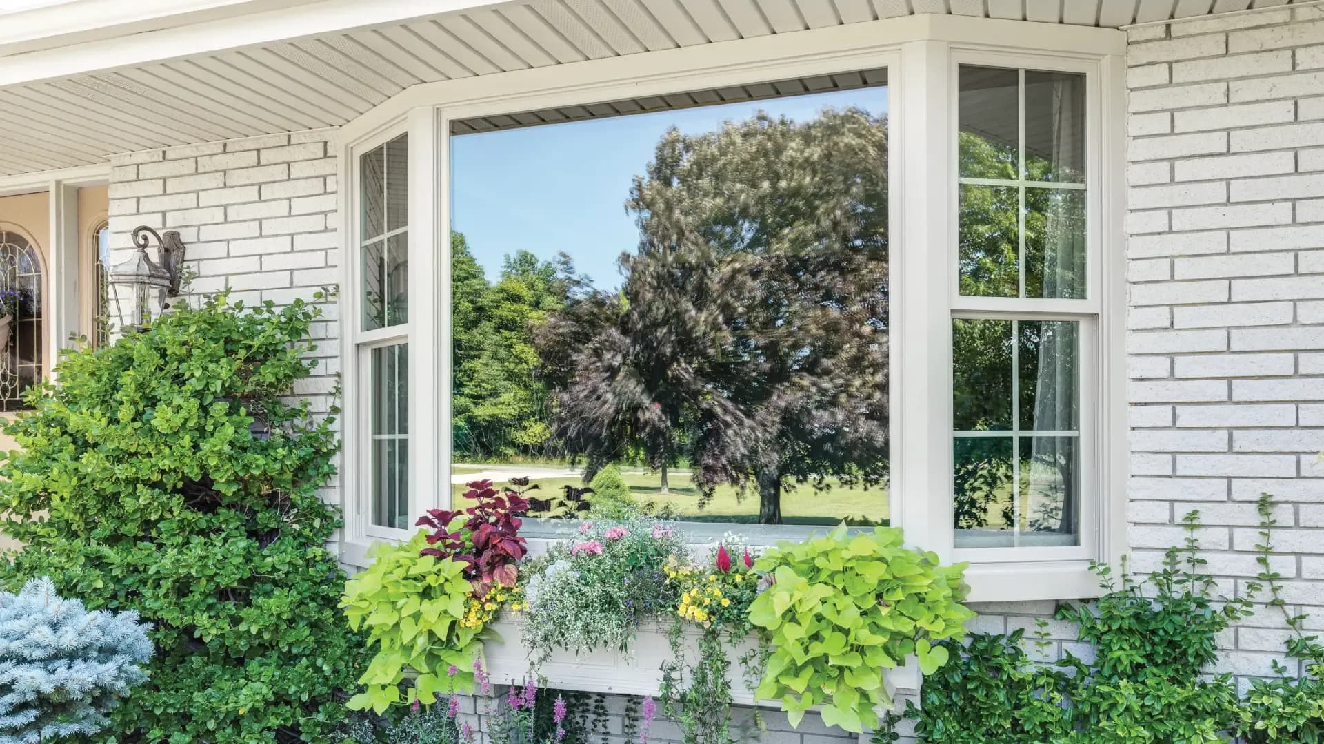 White vinyl bay window with a large central picture pane and double-hung side windows with grilles, installed on a light grey painted brick house surrounded by colorful landscaping.