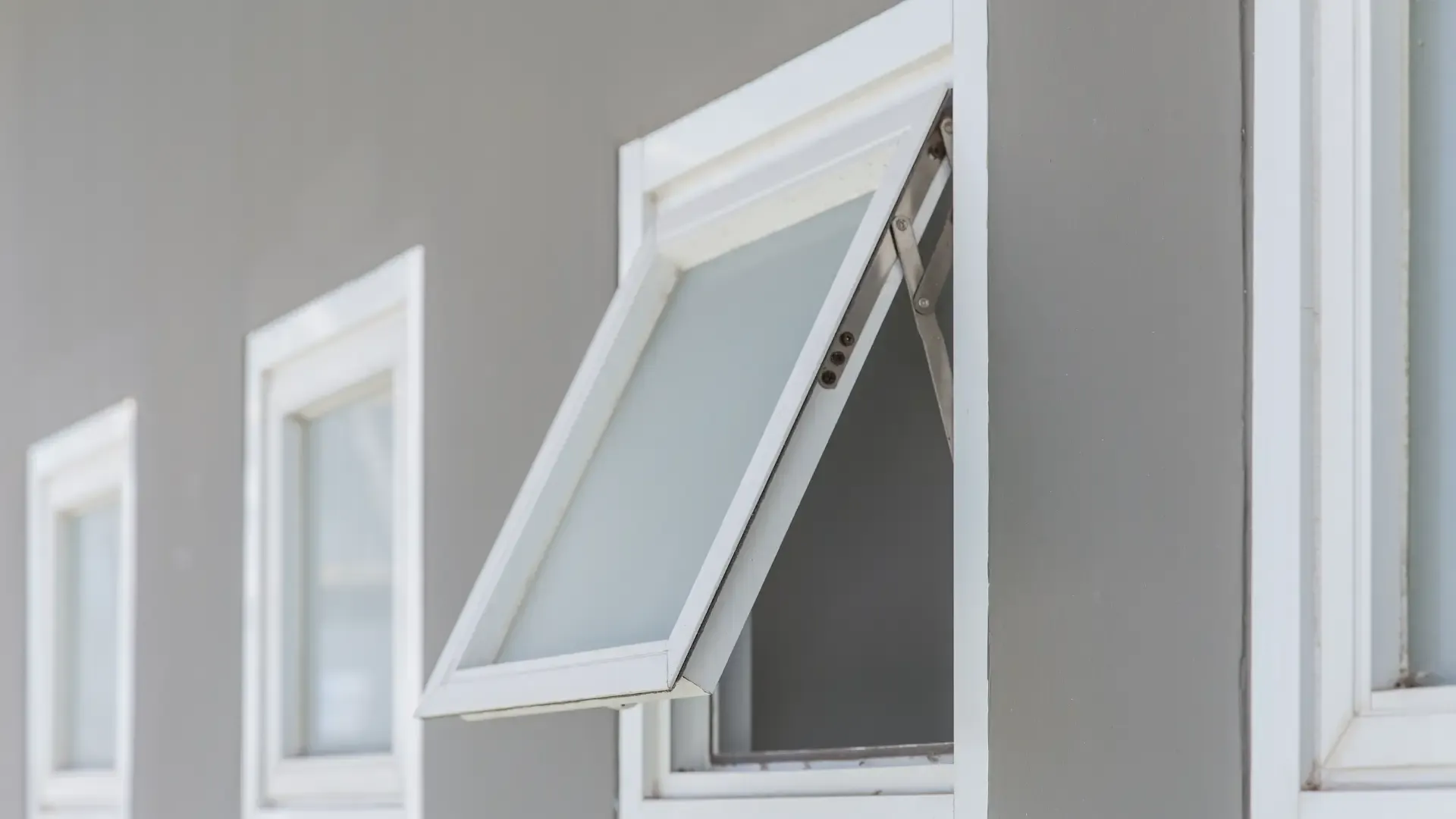 Side view of an open white awning window with frosted glass set in a light grey exterior wall.