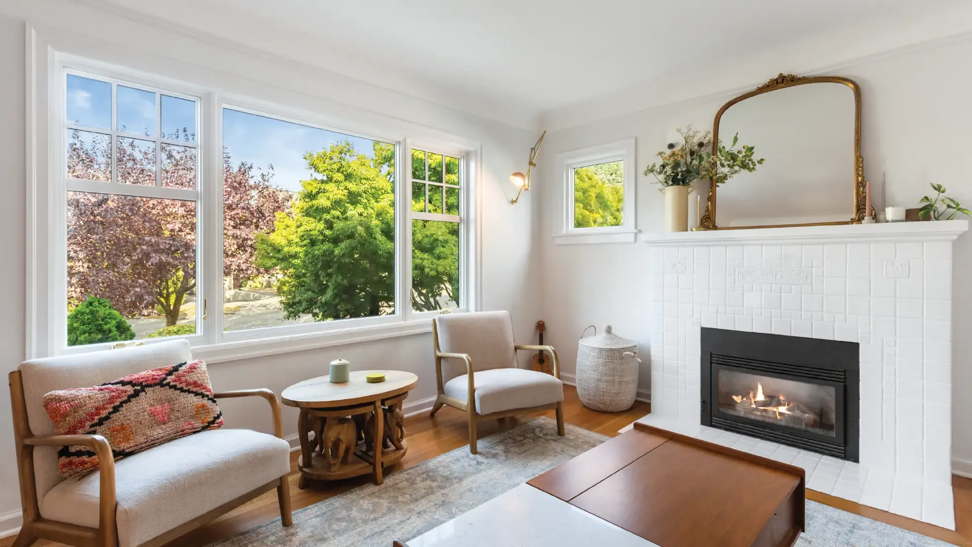 Brightly lit living room featuring large white-framed windows with top grilles, neutral armchairs, and a white tiled fireplace with a gold mirror above it.