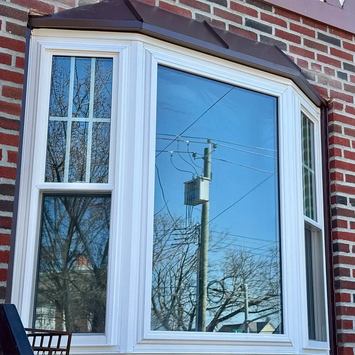 White vinyl bay window with a brown metal roof overhang, installed on a red brick exterior wall with sky and telephone pole reflections.