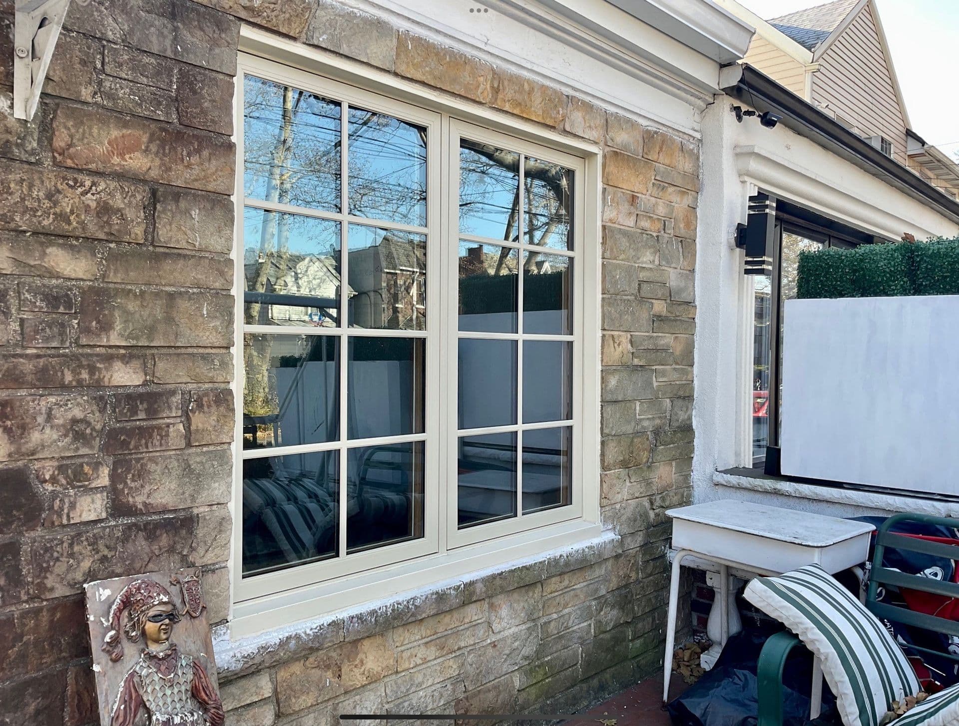 Exterior view of a beige-framed double-hung window unit with colonial grilles, installed into a rustic stone block wall above a patio setup with outdoor furniture.