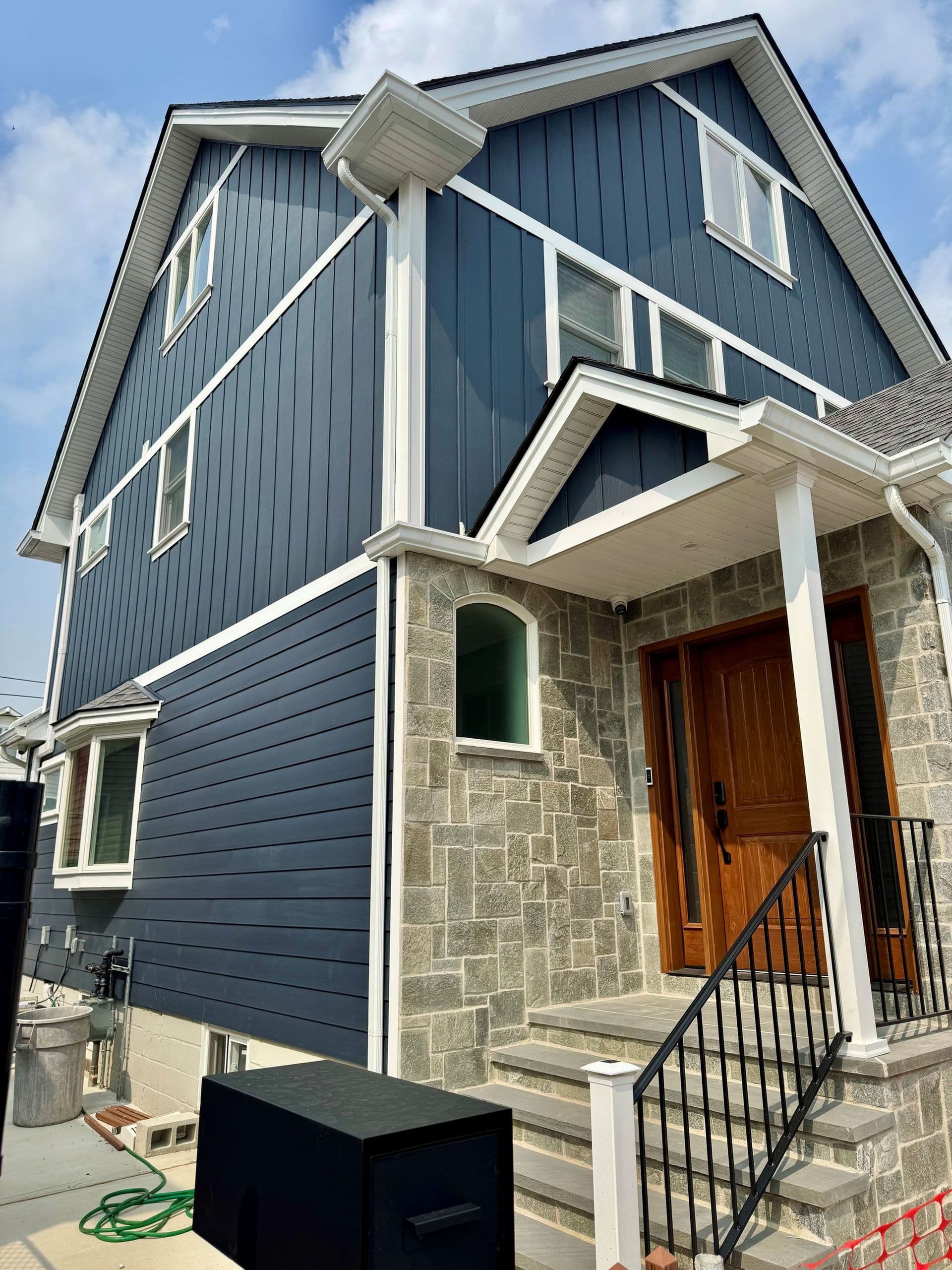 Angled exterior view of a multi-story home featuring dark navy blue siding—vertical board-and-batten on the upper gables and horizontal lap siding on the lower level—accented with crisp white trim, a stone veneer entryway portico with a wood front door, and a black metal handrail leading up the front steps.