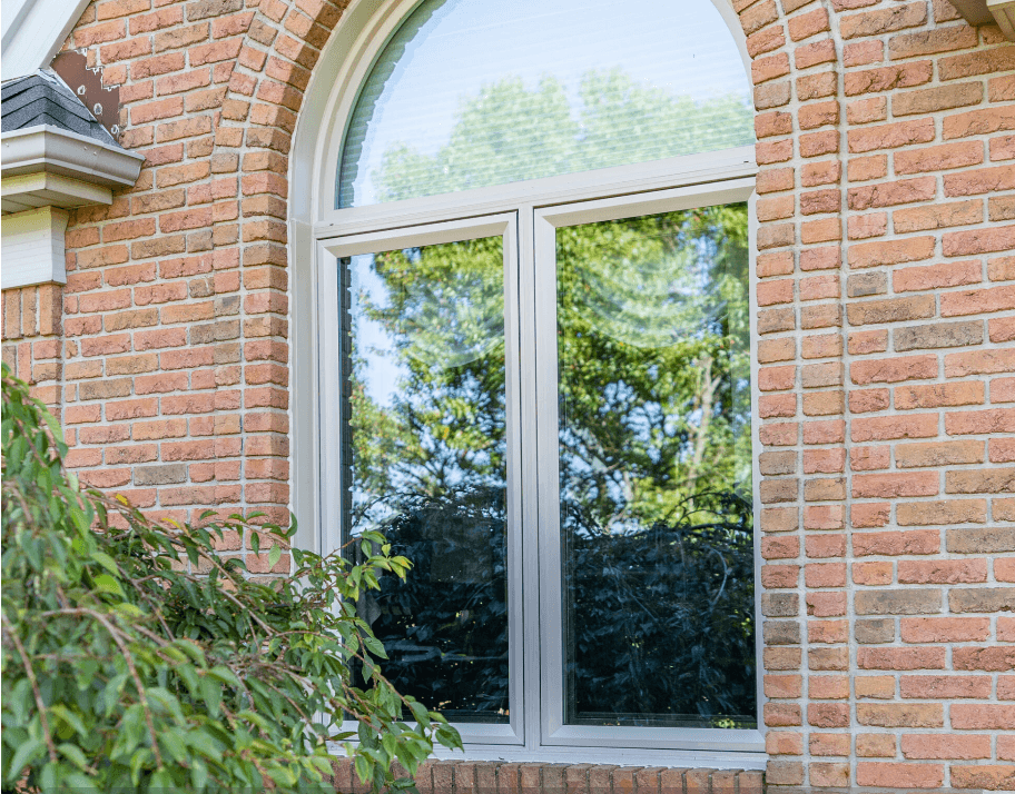 Arched window with white frame set in a brick wall reflects lush green trees. Nearby leafy bush adds a natural feel to the scene.