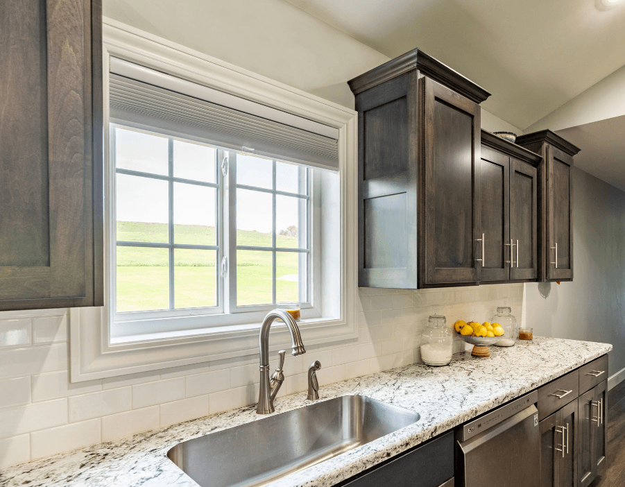 Interior view of a kitchen featuring a white-framed casement window with colonial grilles positioned above an stainless steel undermount sink. The kitchen includes dark wood cabinetry, white subway tile backsplash, and speckled granite countertops.