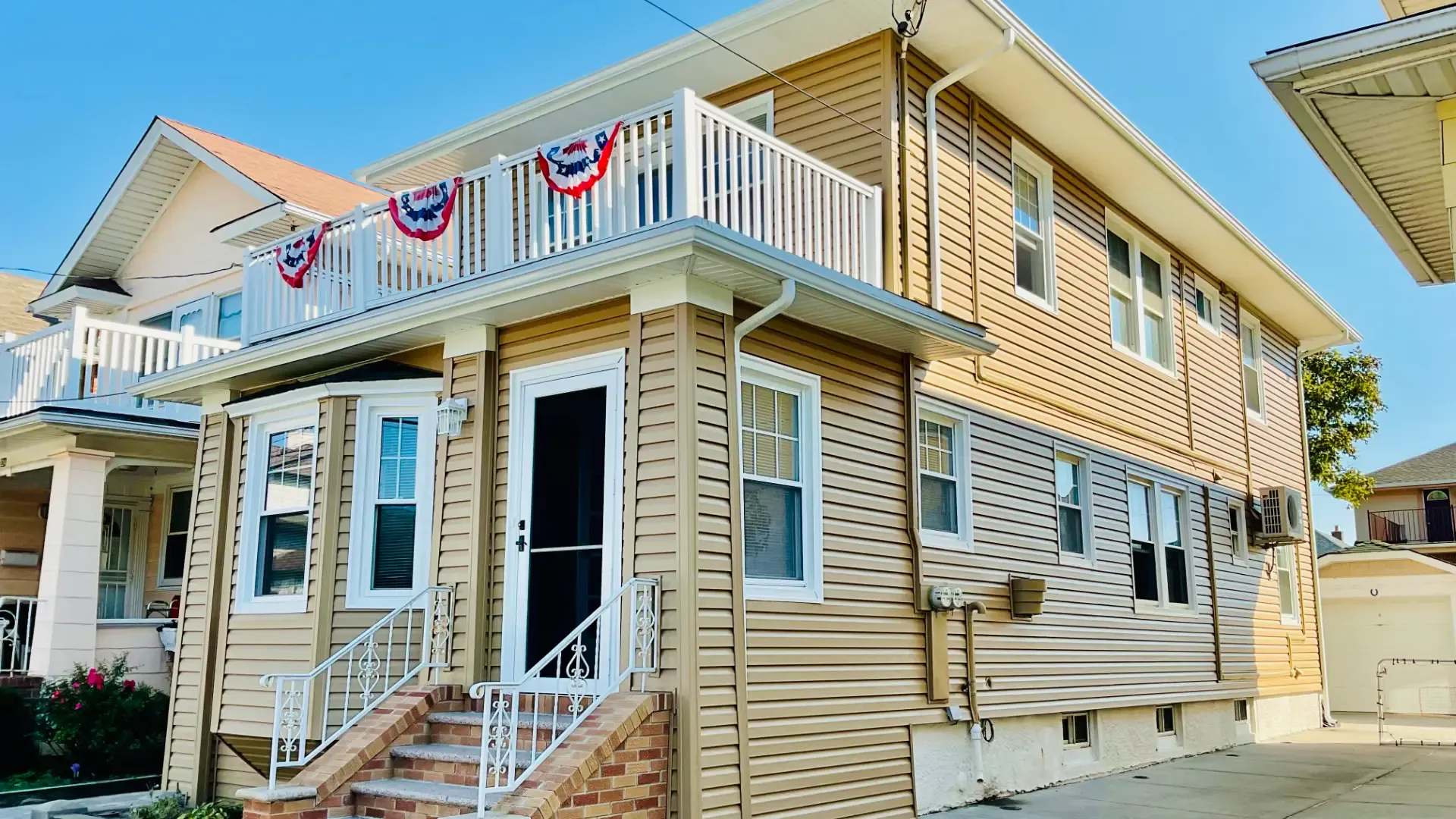Two-story tan vinyl-sided house featuring a second-floor balcony decorated with patriotic bunting and white railing.