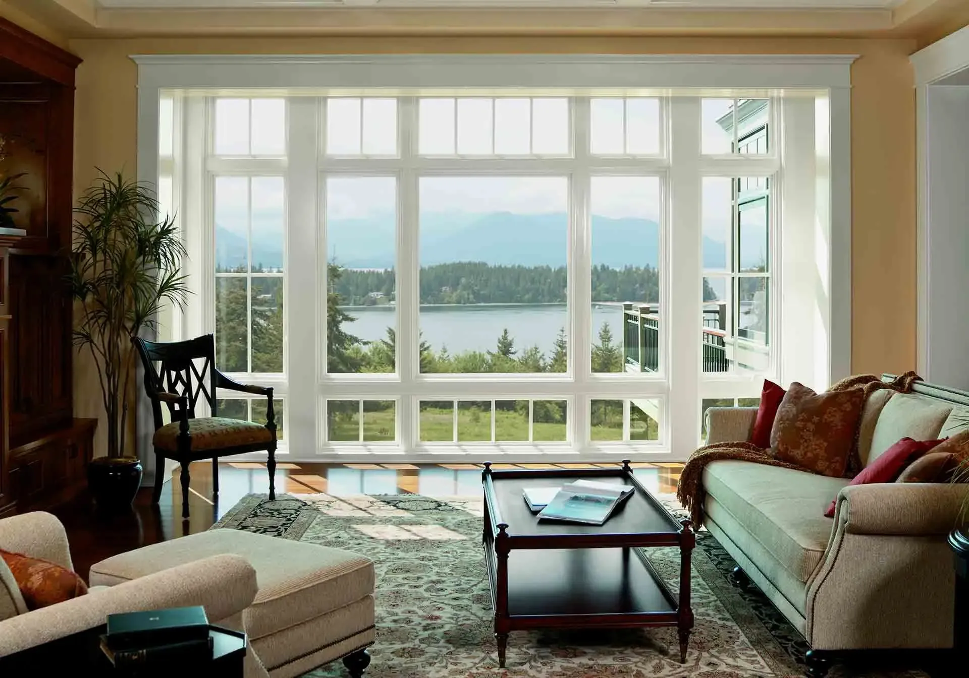 An interior view of a bright, open living room with a expansive wall of white-framed window combinations. The large window wall consists of clear picture windows surrounded by upper transoms and lower accents with grilles, offering a wide scenic view of a lake and surrounding forest. The room is decorated with a sofa, an armchair, an ottoman, a coffee table, and warm hardwood flooring.
