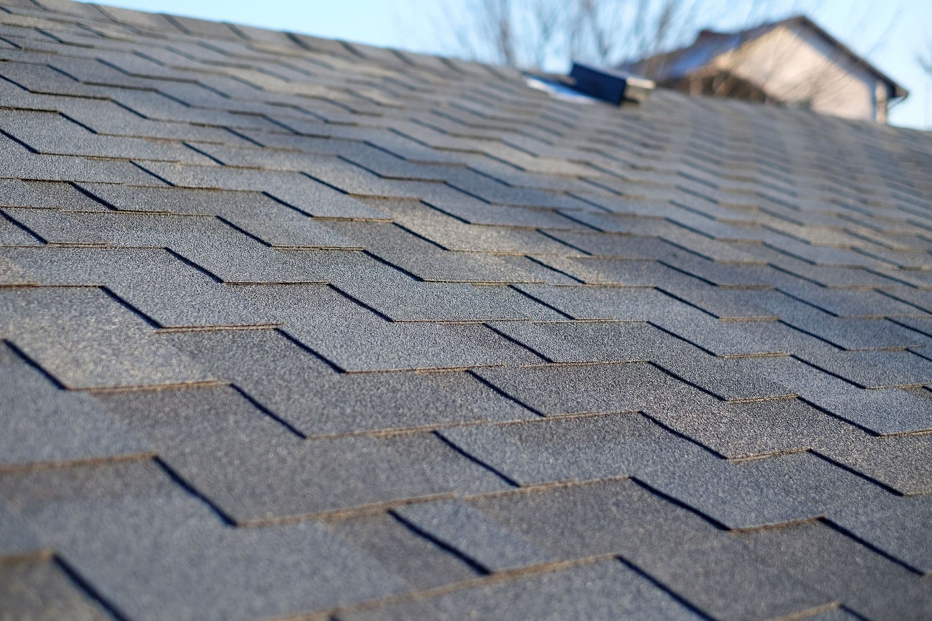 A close-up perspective view of dark grey bitumen asphalt roof shingles featuring a geometric honeycomb or hexagonal cut pattern installed along a roof slope.