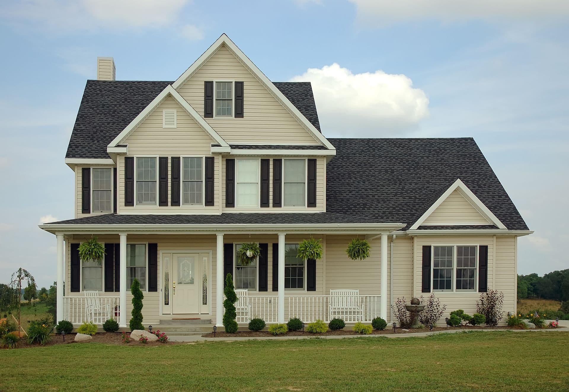 Two-Story Beige Farmhouse with Covered Porch (bigstock-Farm-House-150703 (1).jpg) Front exterior view of a two-story beige siding farmhouse featuring a black shingle roof, dark brown or black window shutters, a white front door with sidelights, a prominent covered front porch with white railings and hanging plants, and a manicured green lawn under a lightly clouded sky.