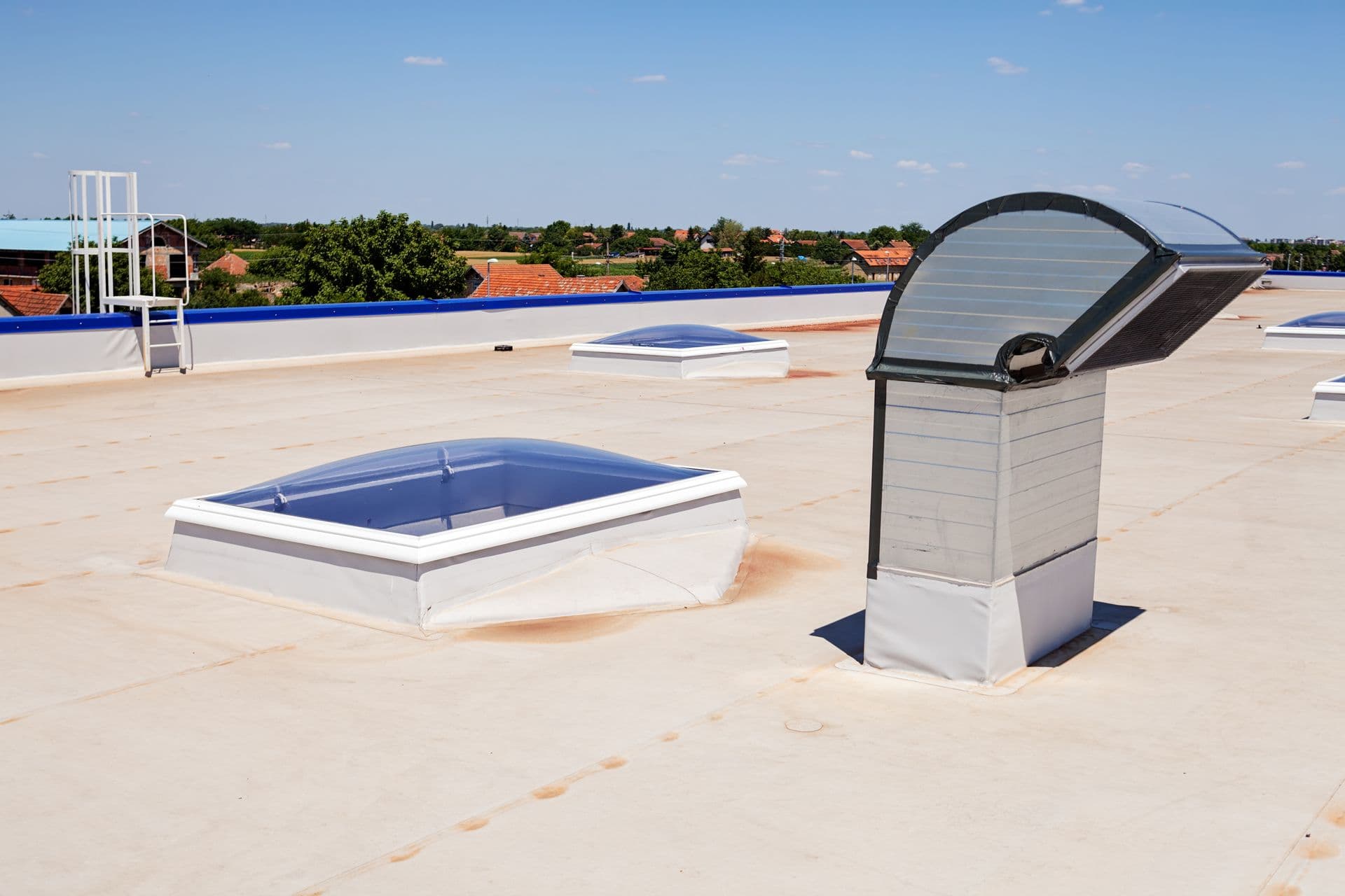 Wide perspective view of an expansive white membrane flat roof on an industrial building, featuring raised domed skylights, a gooseneck ventilation exhaust hood, and a blue perimeter parapet wall under a clear blue sky.