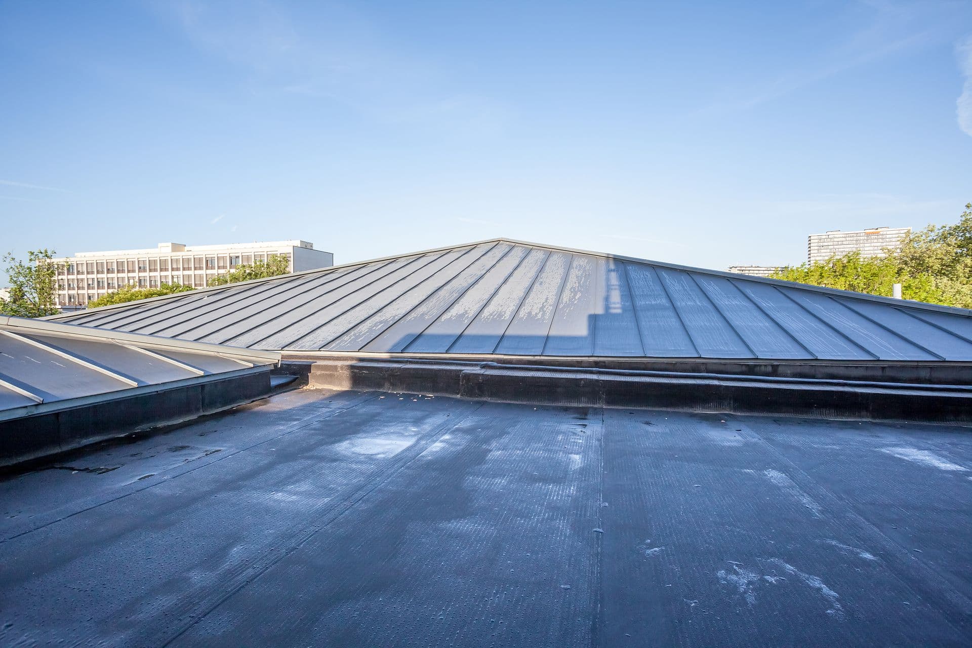 A wide perspective view looking over a dark grey flat rubberized roof section toward a low-sloped, grey standing seam metal roof structure under a clear blue sky, with urban buildings visible in the distance.