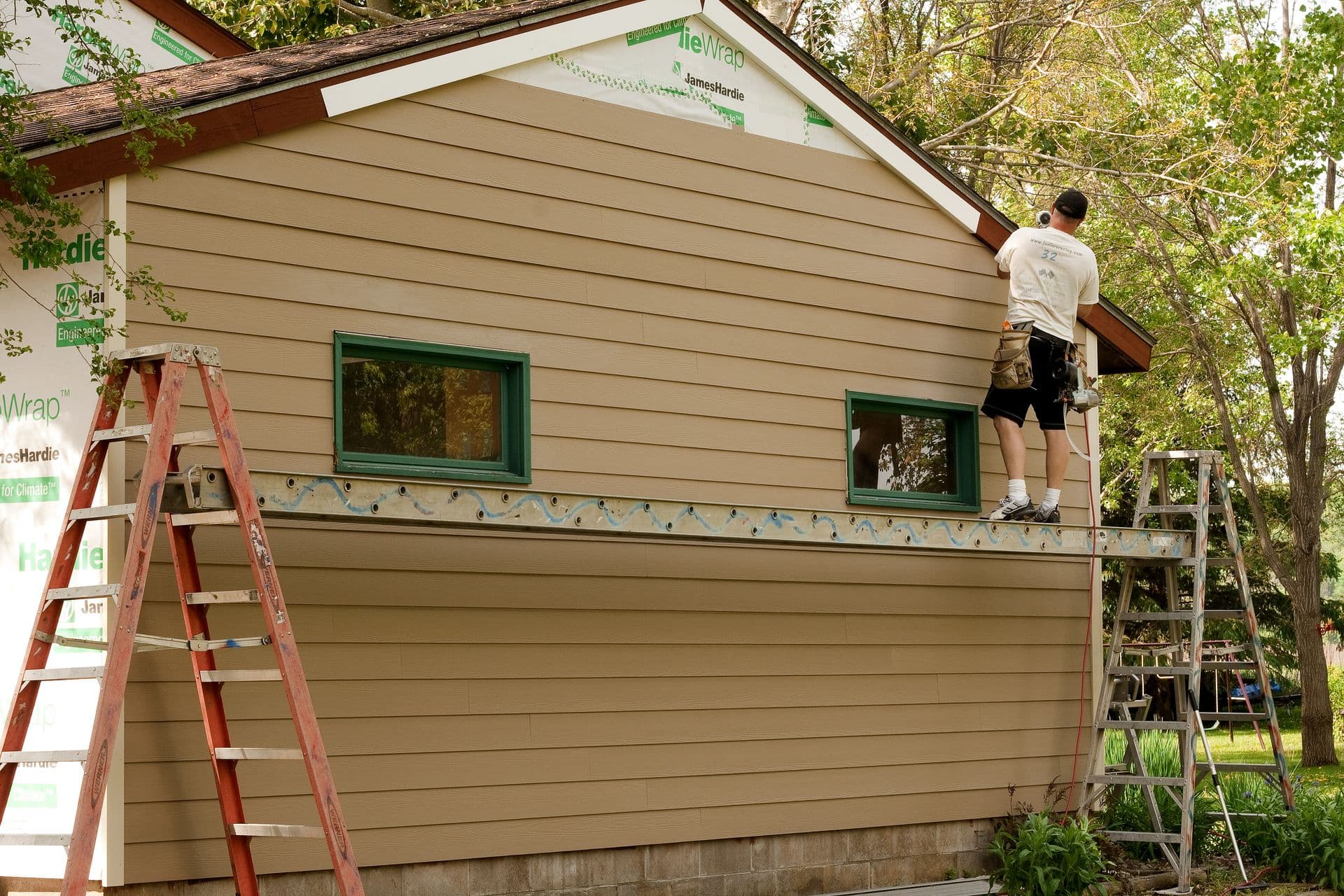 A contractor wearing a tool belt stands on an elevated plank scaffolding supported by two stepladders, installing tan James Hardie fiber cement lap siding onto the exterior gable wall of a house featuring green-framed windows and visible house wrap.