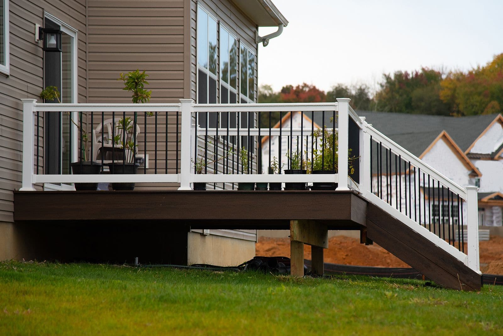 Side exterior view of an elevated dark brown composite deck attached to a house with beige vinyl siding, featuring a white vinyl railing system with black metal balusters, potted plants along the deck edge, and staircase steps leading down to the lawn.