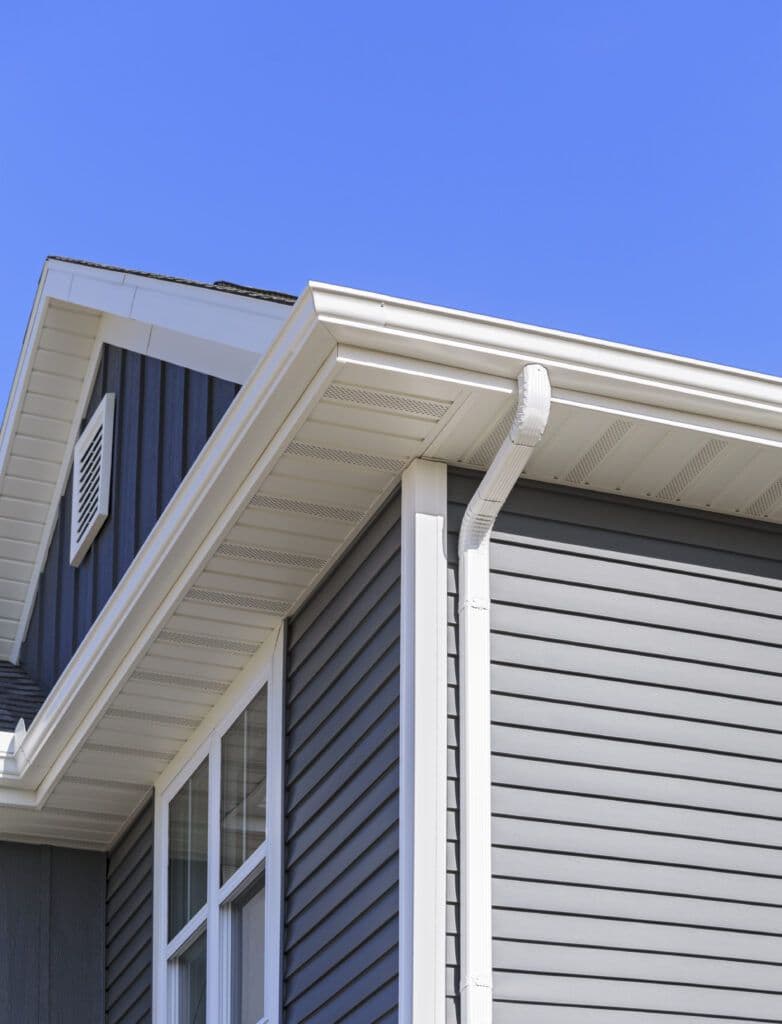 Modern residential corner detail showing white gutters, downspout, soffit vents, and gray horizontal siding against clear blue sky