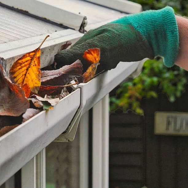 Person wearing green gloves cleaning autumn leaves and debris from a roof gutter