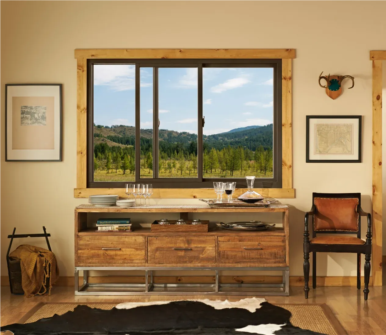 Dark-framed horizontal sliding window with natural wood trim overlooking a scenic pine forest and rolling hills, set in a rustic room above a wooden sideboard table.