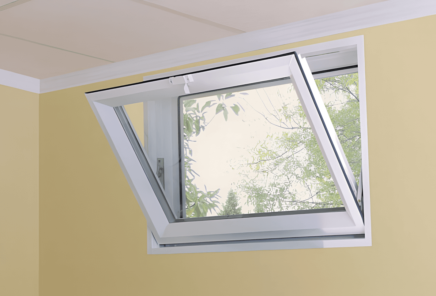 Close-up interior view of a white vinyl hopper window tilted inward from the top, set into a light yellow wall and looking out toward green trees.